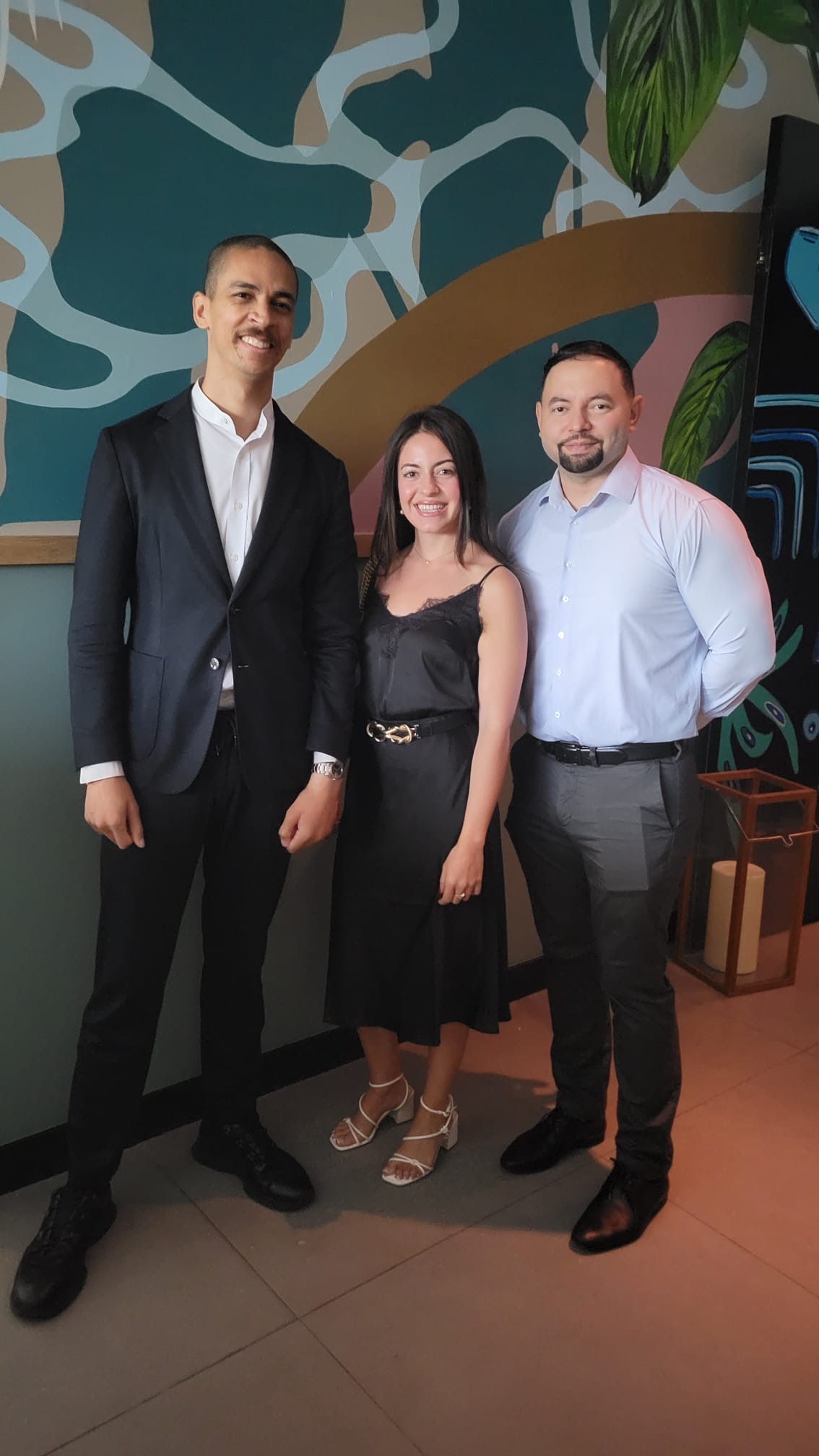 Three people, two men and one woman, are standing indoors against a colorfully patterned wall. One man is in a suit, the woman in a black dress, and the other man in a light blue shirt and dark pants.