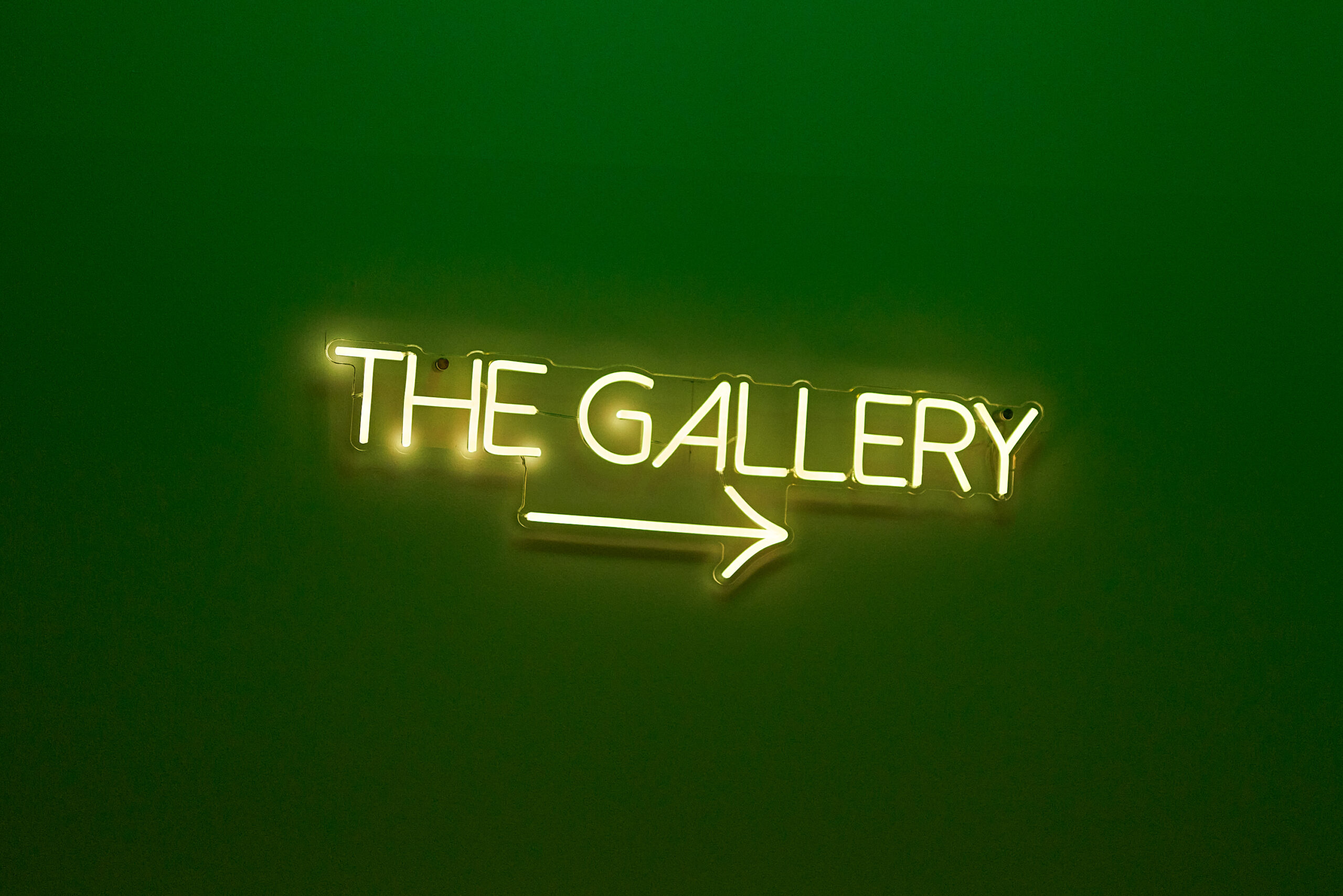 A neon sign with the words "THE GALLERY" and an arrow pointing to the right, glowing brightly against a dark green background.