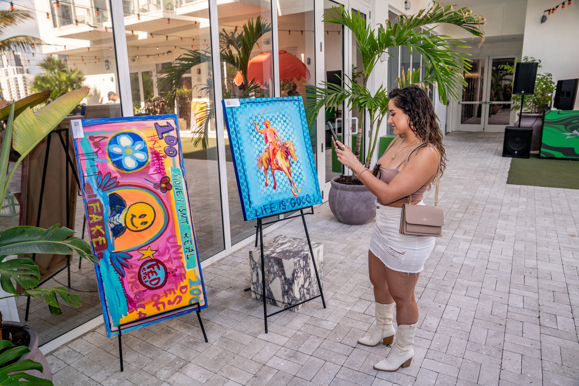 A woman in a white dress and boots takes a photo of two colorful paintings displayed on easels in an outdoor setting with potted plants around.