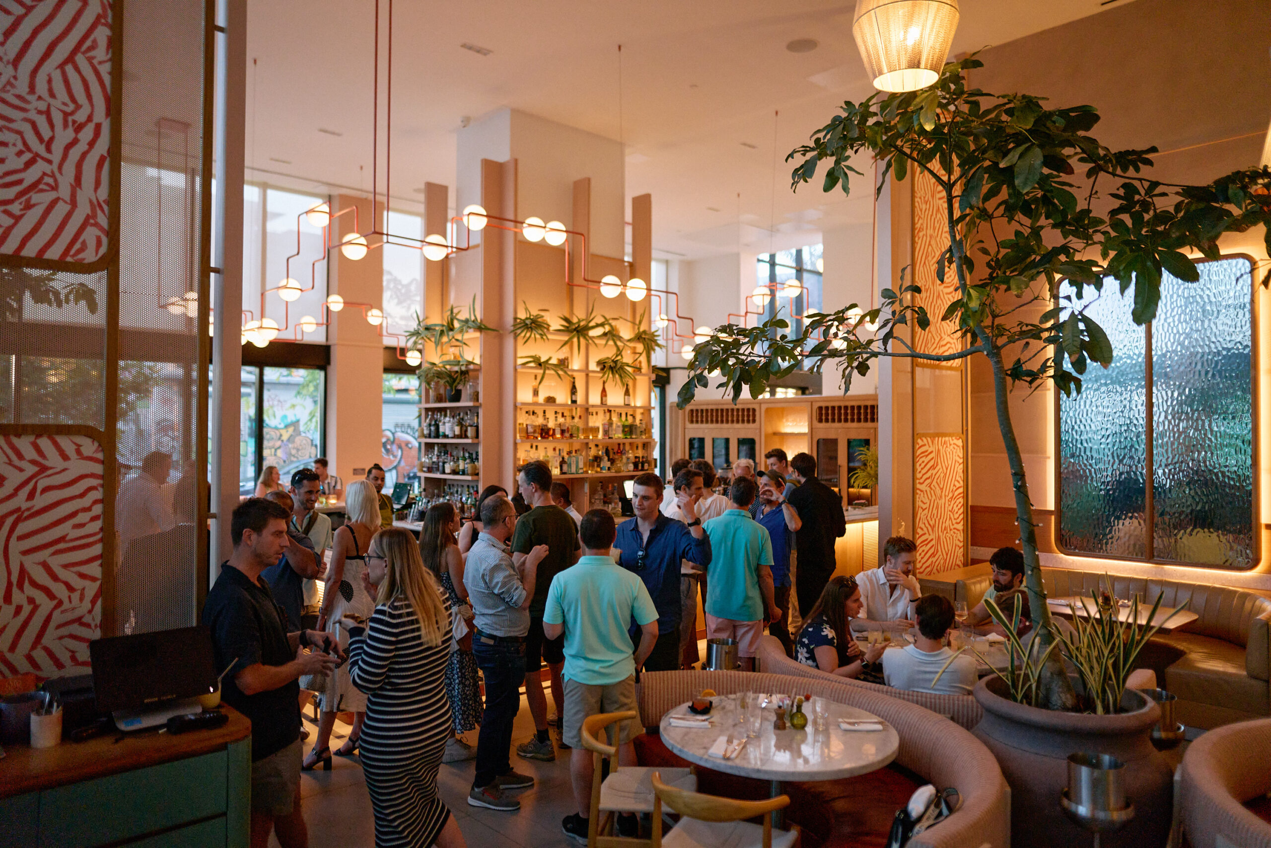 A group of people socializing in a modern bar with green plants, large windows, and ambient lighting. Some are seated while others stand near the bar area.