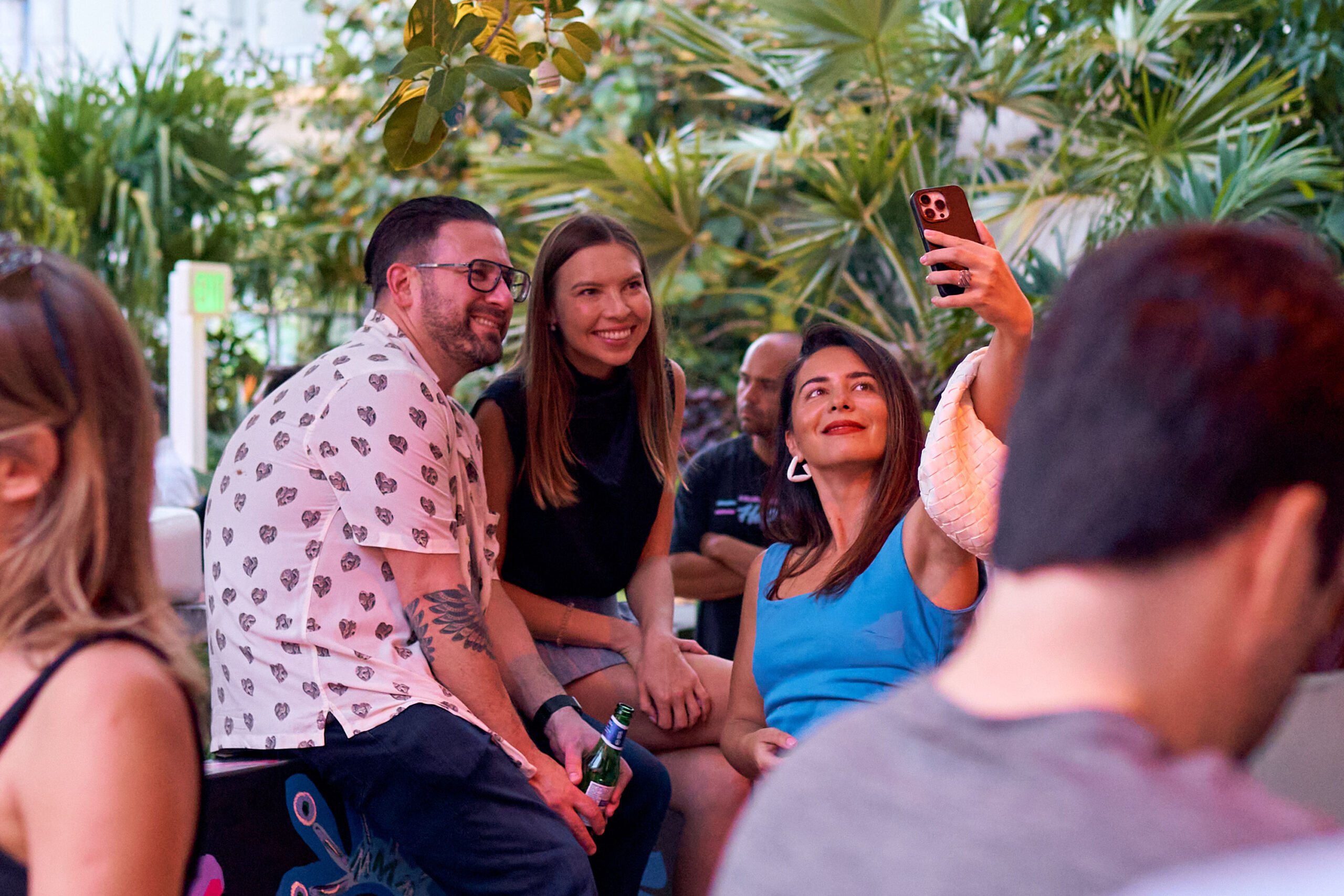 Three people are posing for a selfie outdoors, surrounded by greenery. One person is holding a phone to take the photo, while the others sit close and smile.