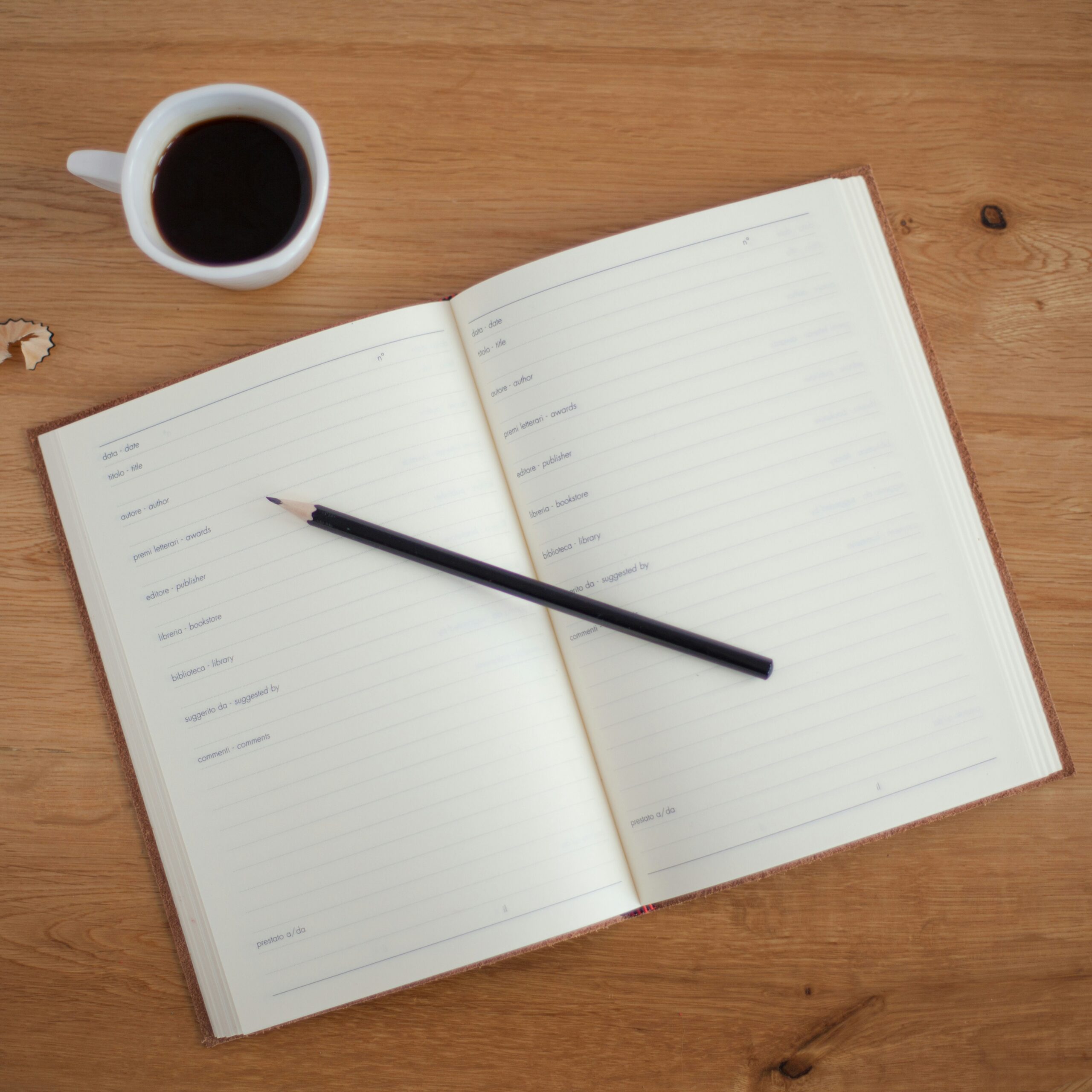 An open lined notebook with a pencil placed diagonally across it on a wooden table. A cup of black coffee is on the top left of the notebook.