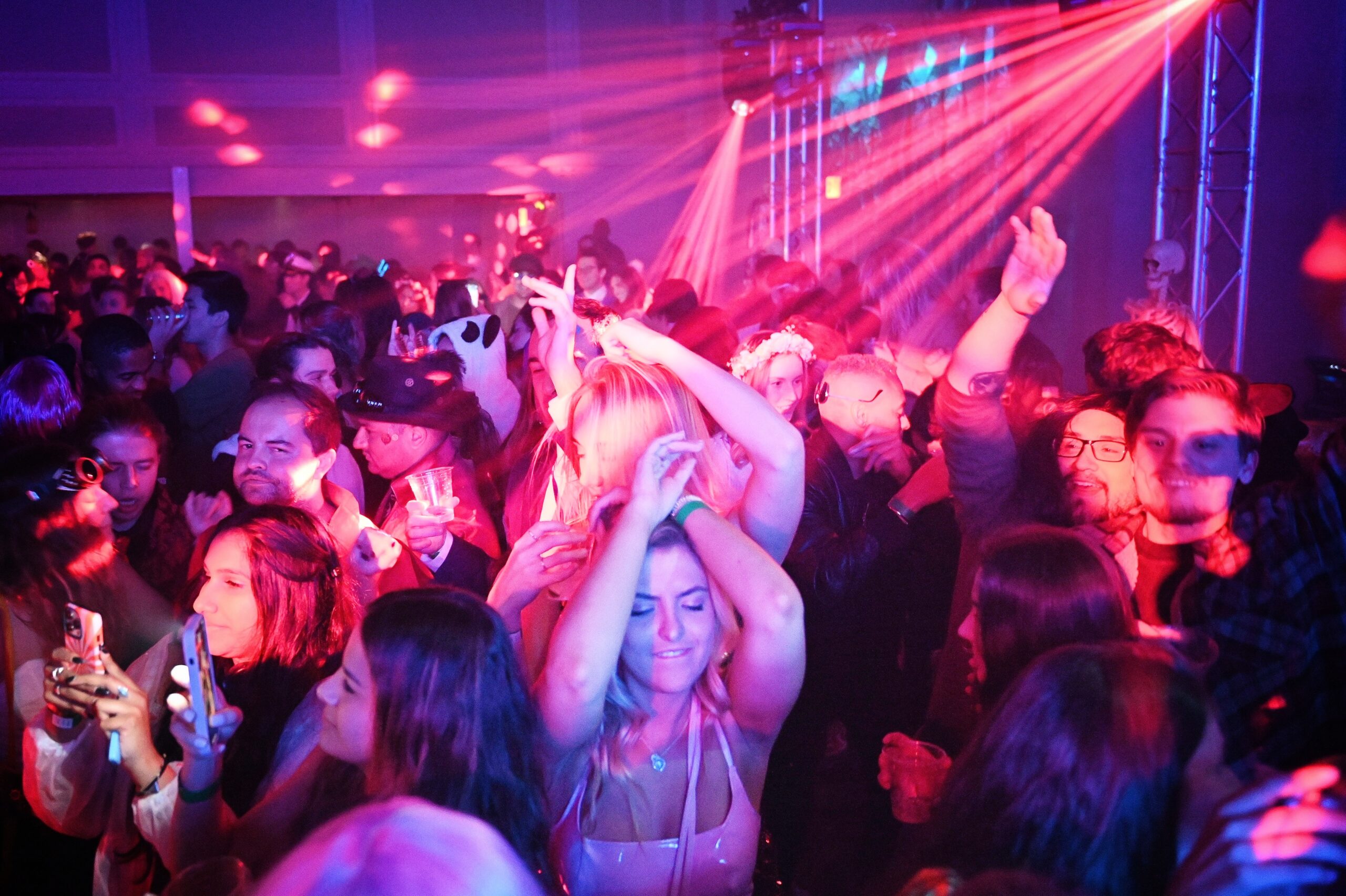 A crowded dance floor with people dancing and enjoying a colorful light show. Some individuals are holding drinks, and a few are taking photos with their phones.