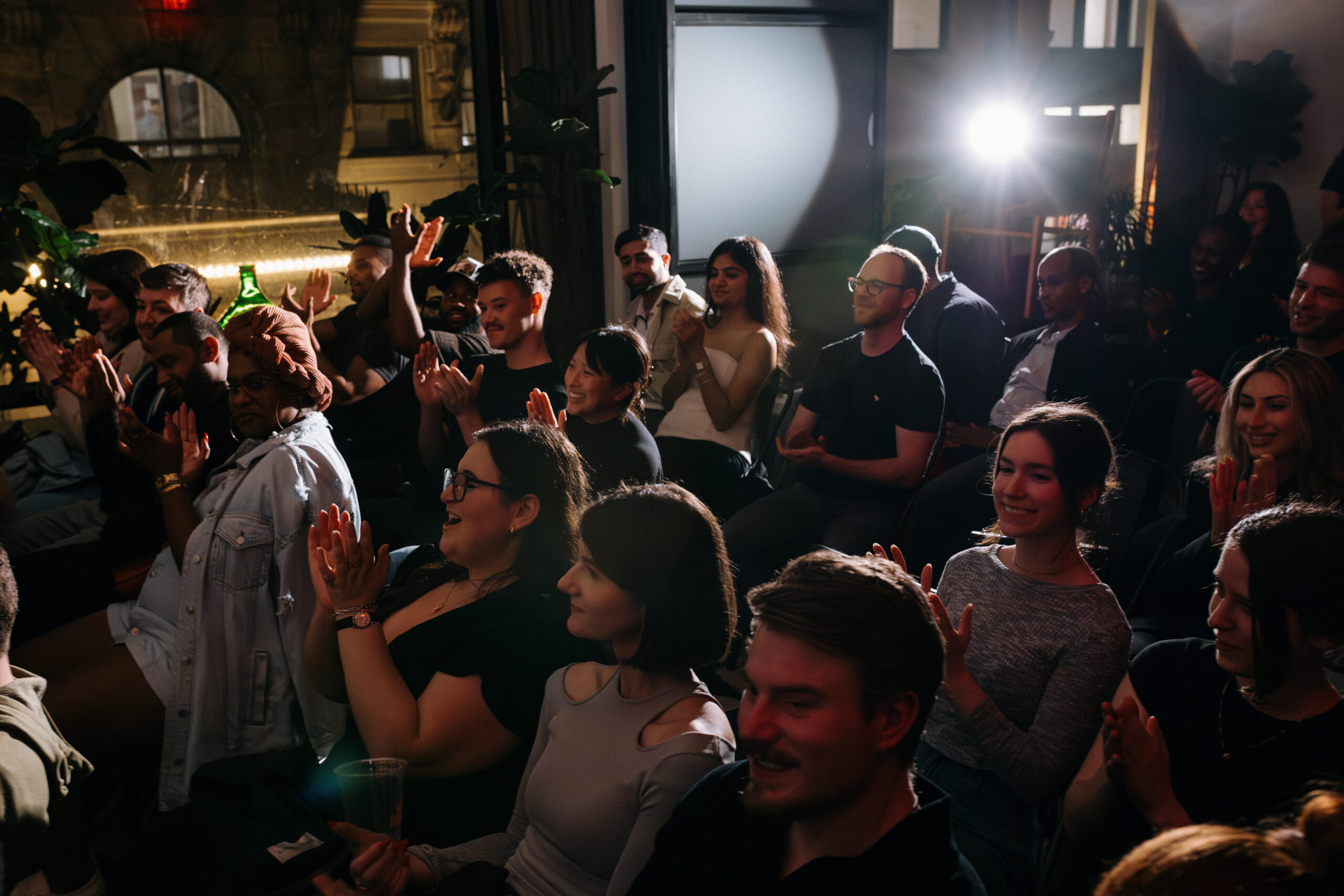 A diverse group of people seated in a dimly lit room, clapping and looking towards the front, suggests they are attending an event or presentation.