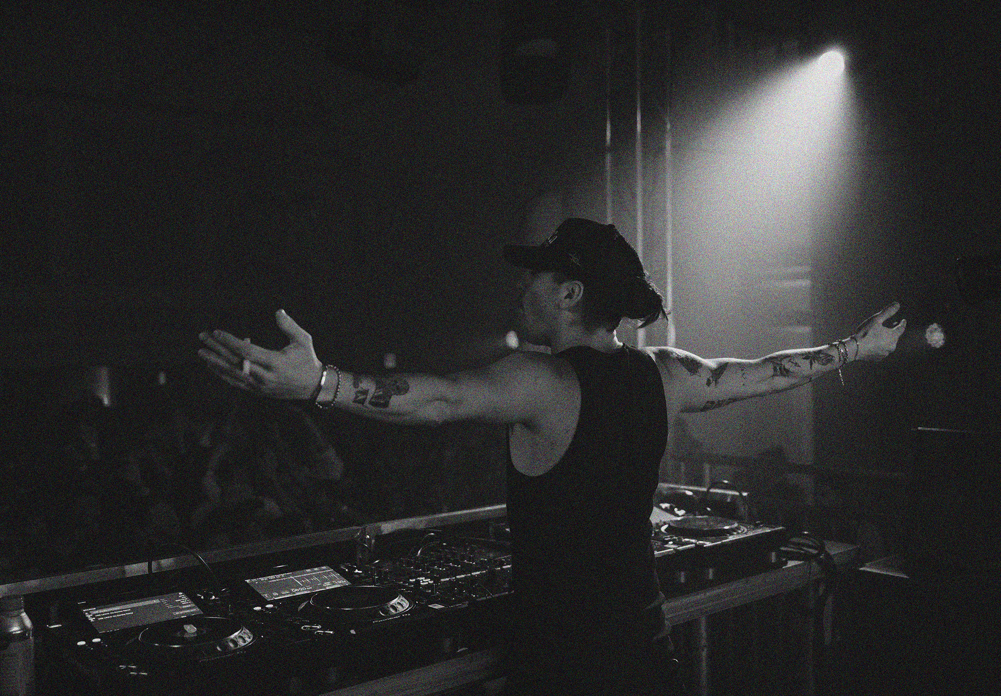 A DJ wearing a tank top and a cap with arms outstretched stands behind a DJ console in a dimly lit club with a crowd in the background.