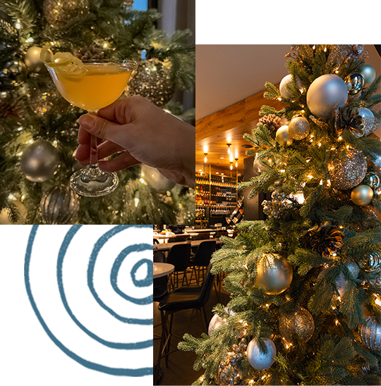 A hand holding a cocktail with a garnish in front of a decorated Christmas tree adorned with silver ornaments and lights. In the background, a restaurant with tables and chairs is seen.