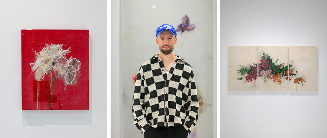 Person standing in front of a gallery wall, featuring a red floral painting to the left and a triptych floral artwork to the right. The person is wearing a checkered shirt and a blue cap.