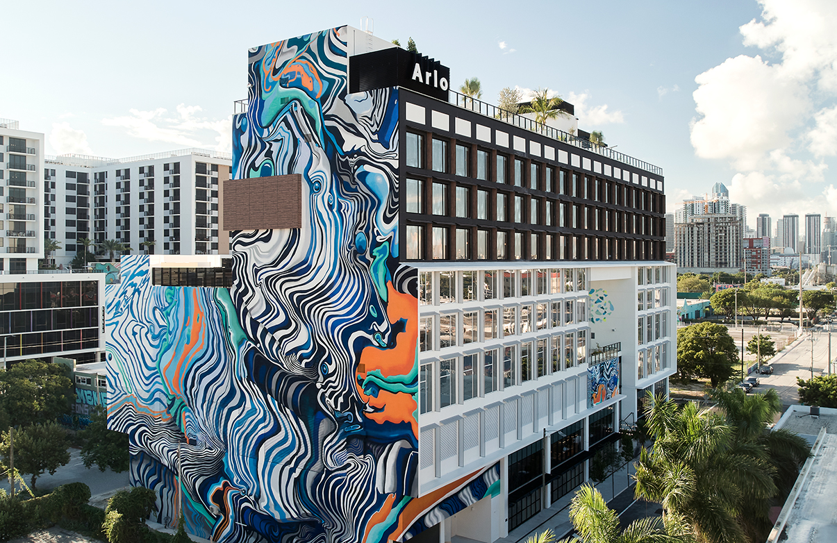 A modern, multi-story building with a vibrant, abstract mural covering its facade. The upper portion has a pattern of windows and a sign with the word "Arlo." Palm trees are visible around the building.