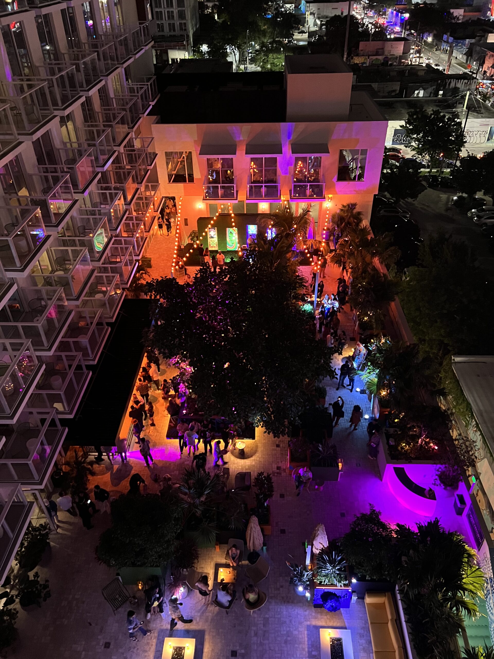 Aerial view of a lively outdoor night gathering with colorful lighting, surrounded by buildings, trees, and people socializing.