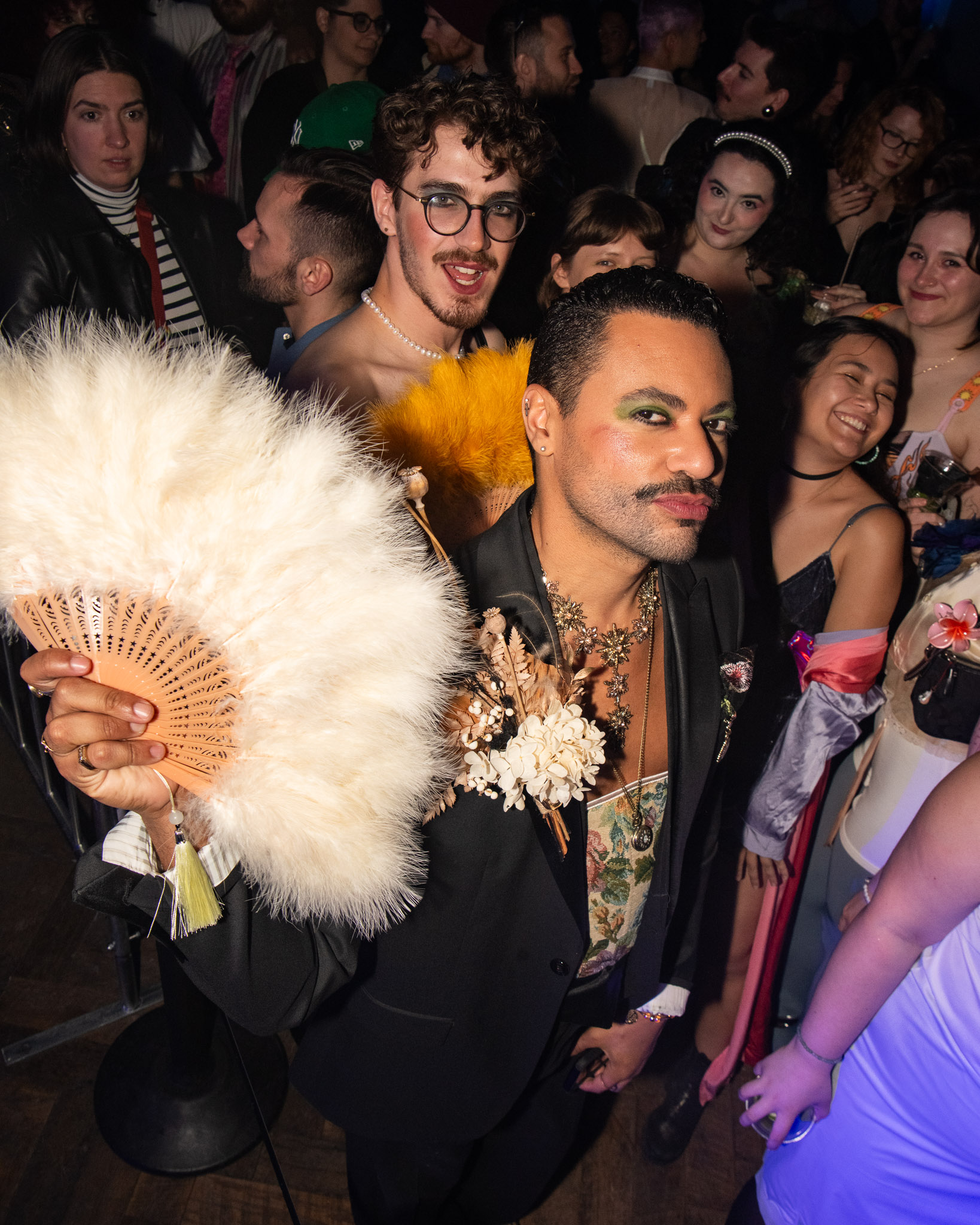 A group of people in a crowded room, with two individuals prominently holding decorative fans. The crowd appears engaged and lively.