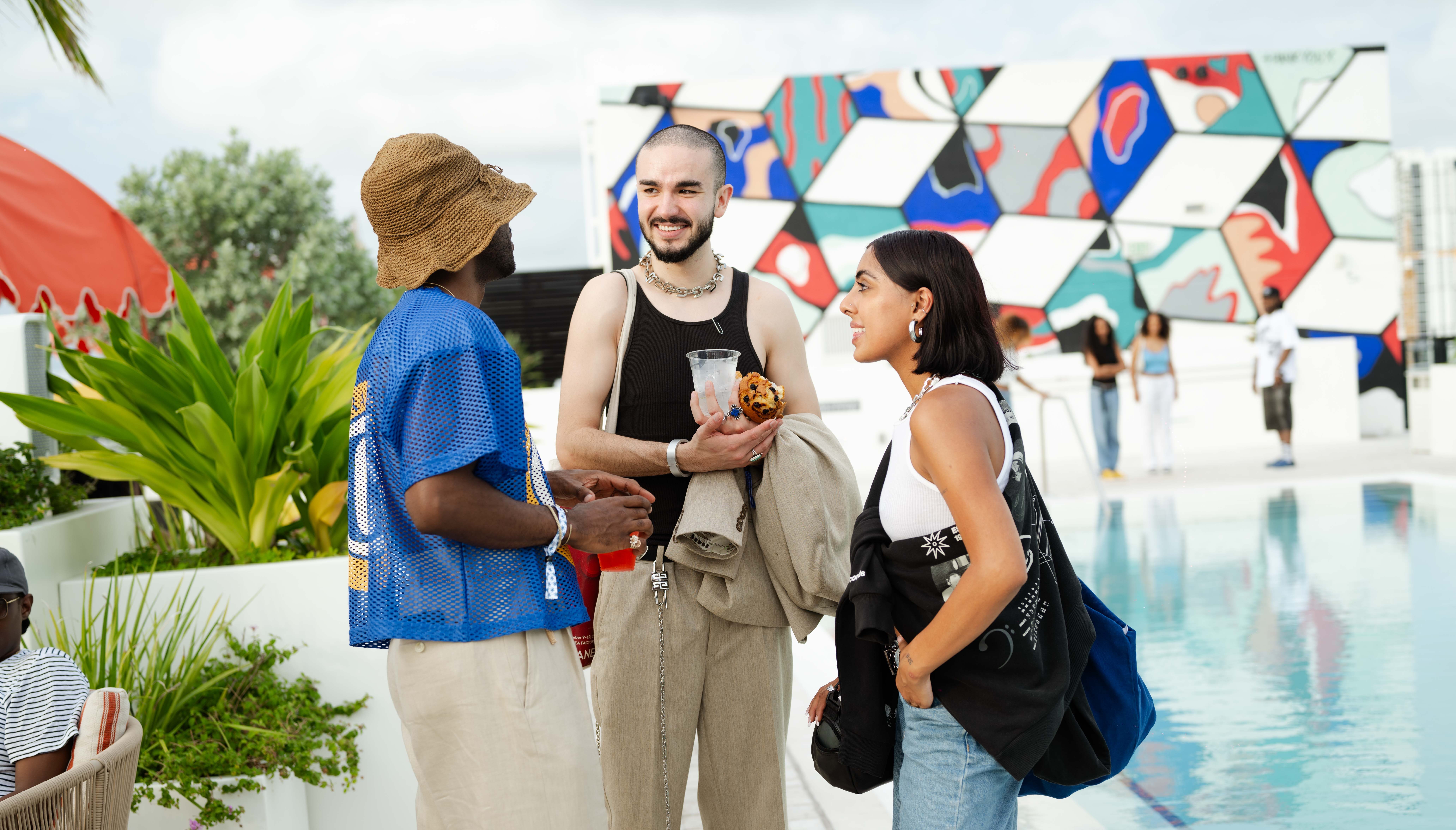 Three people chat by a poolside with geometric murals in the background. Two are holding drinks, and one carries a bag.