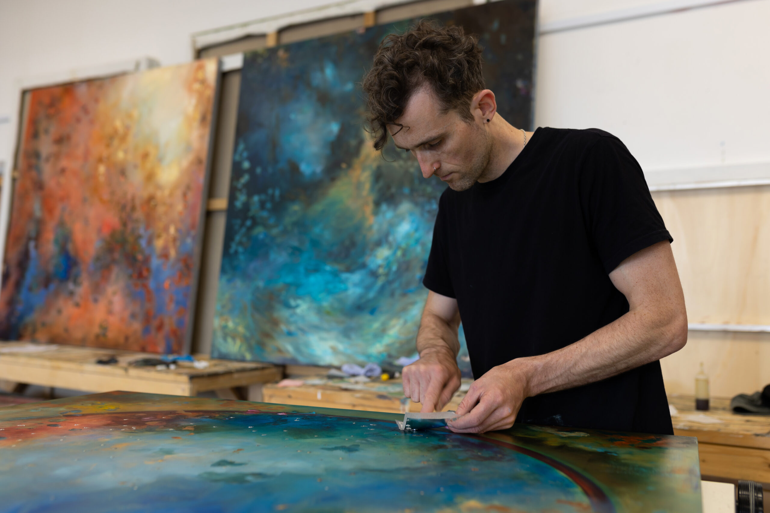 Person working on a large, colorful abstract painting in a studio with similar artworks displayed in the background.