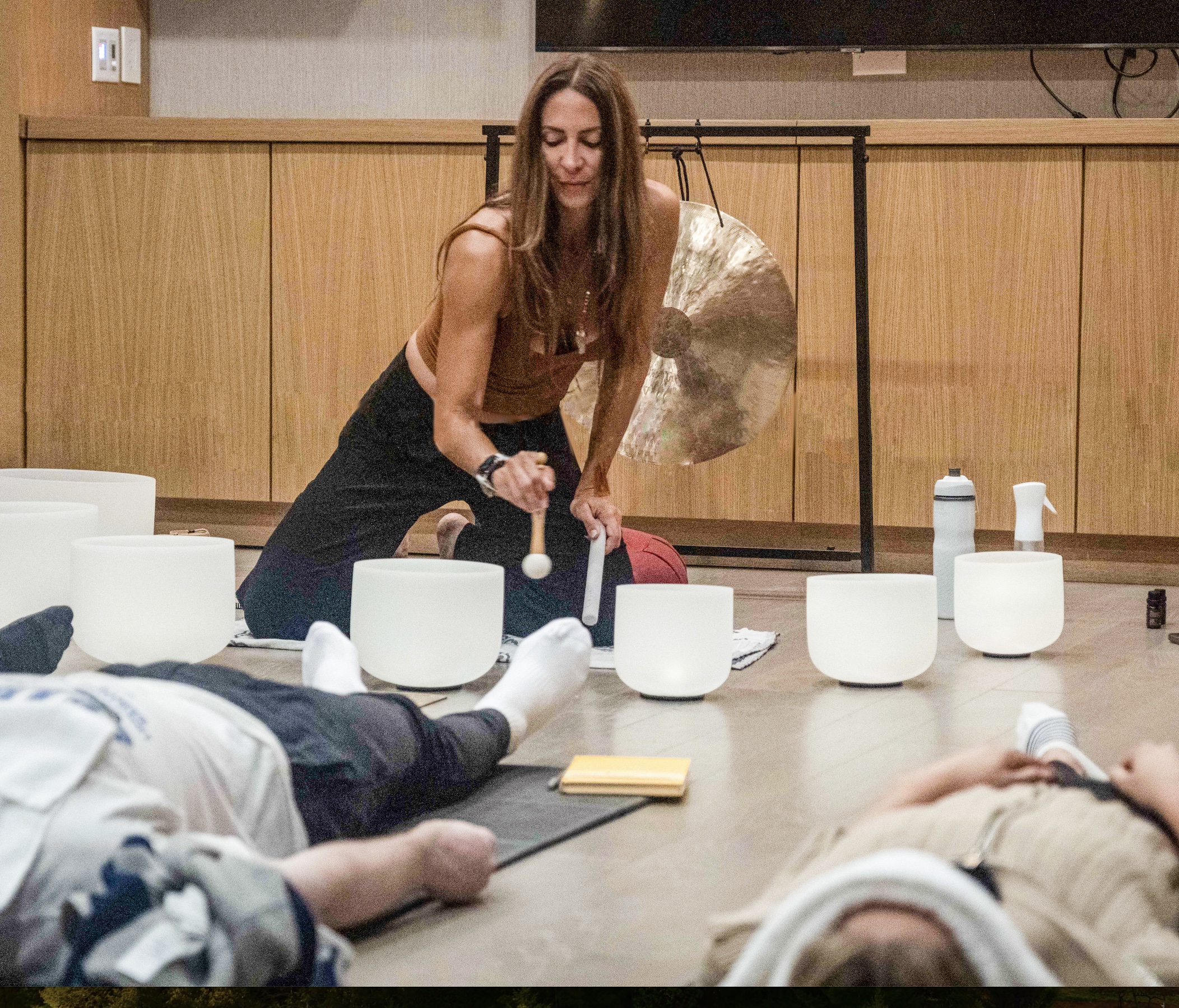 A woman plays crystal singing bowls in a room. Nearby people are lying on the floor, seemingly meditating or relaxing. A gong is visible in the background.