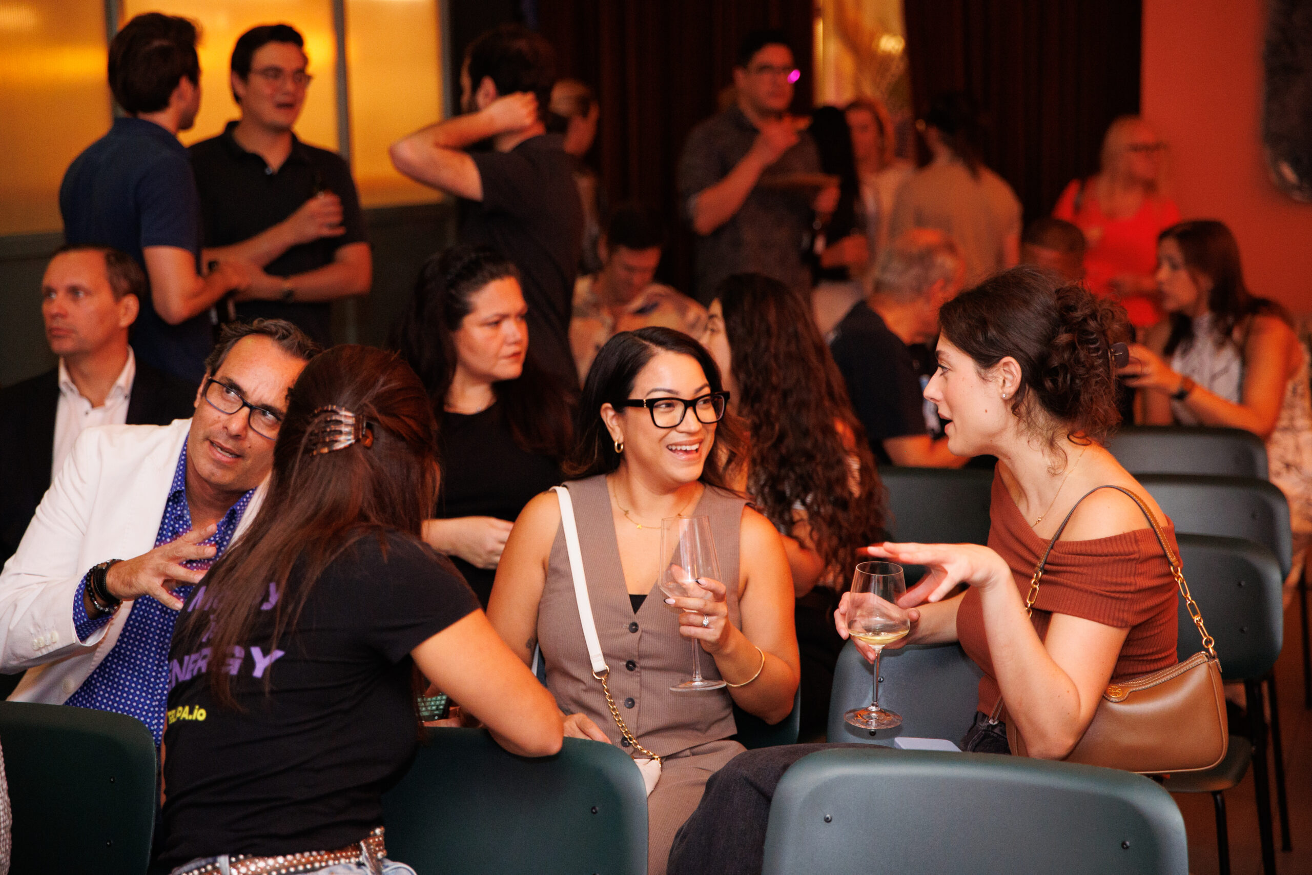 People seated and standing at a social gathering, conversing and holding drinks in a dimly lit room.