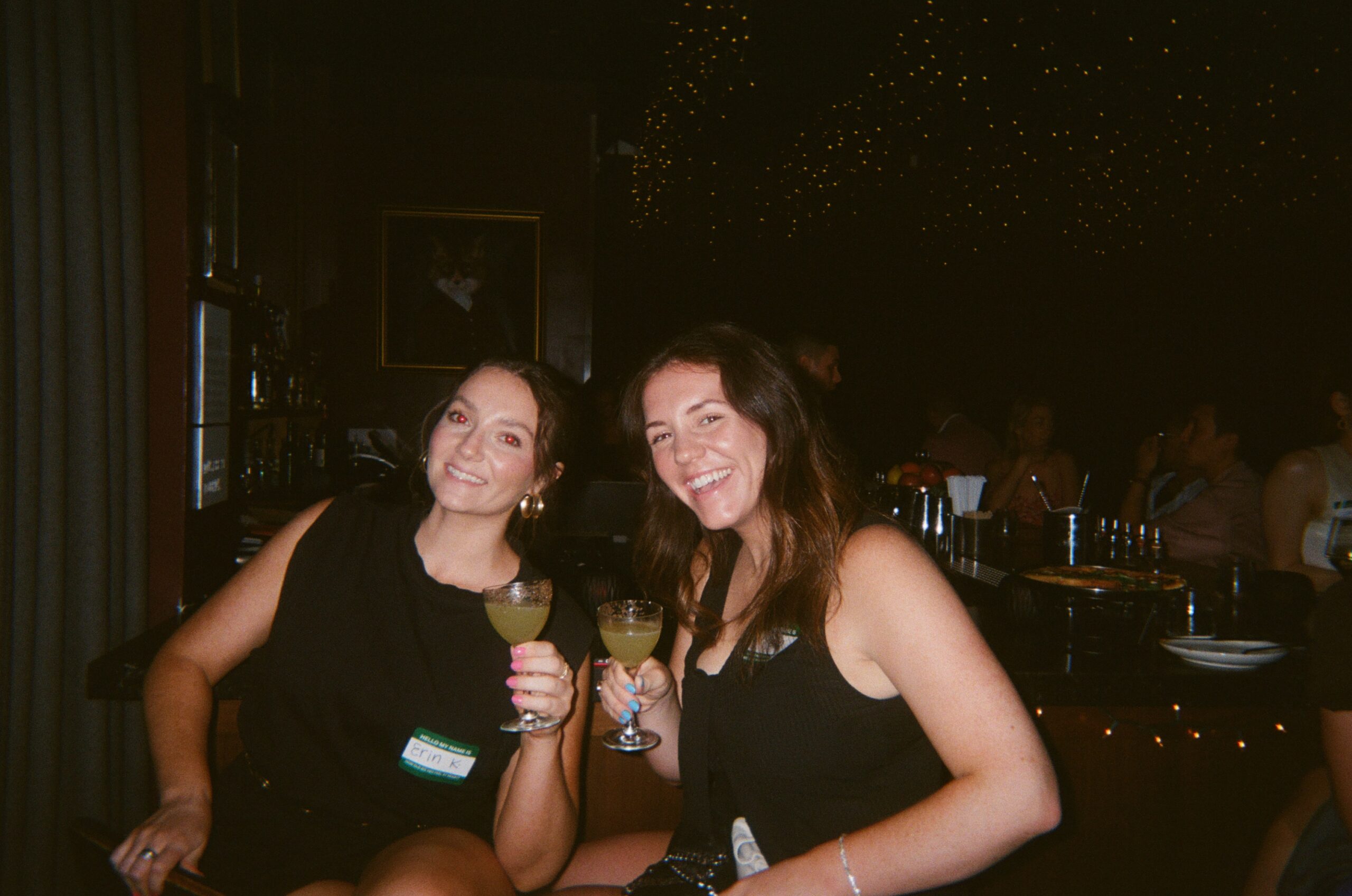 Two people sit at a bar, smiling and holding cocktails. The background features dim lighting and a starry ceiling.