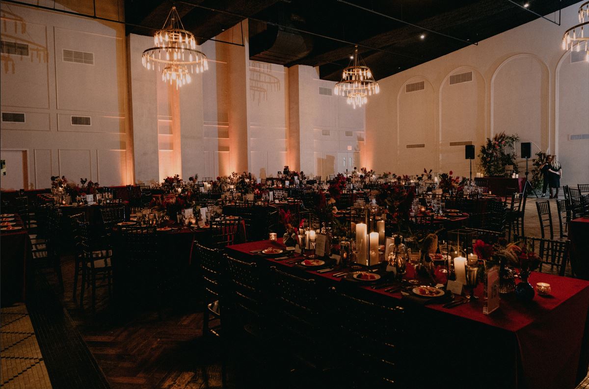 Dimly lit banquet hall with round tables set with candles, plates, and floral centerpieces. Chandeliers hang from the ceiling, casting a warm glow over the room.