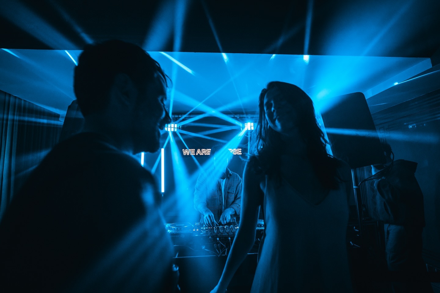 People dancing in a dimly lit room with blue lights and a DJ playing music nearby.