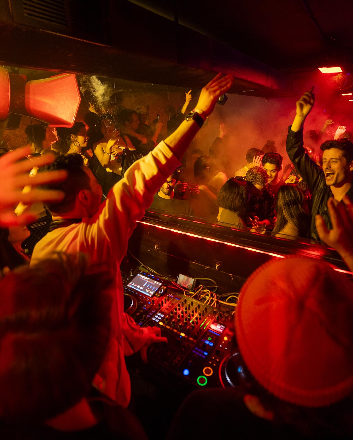 DJ performing in a crowded nightclub with vibrant red lighting; people dancing and raising their hands.