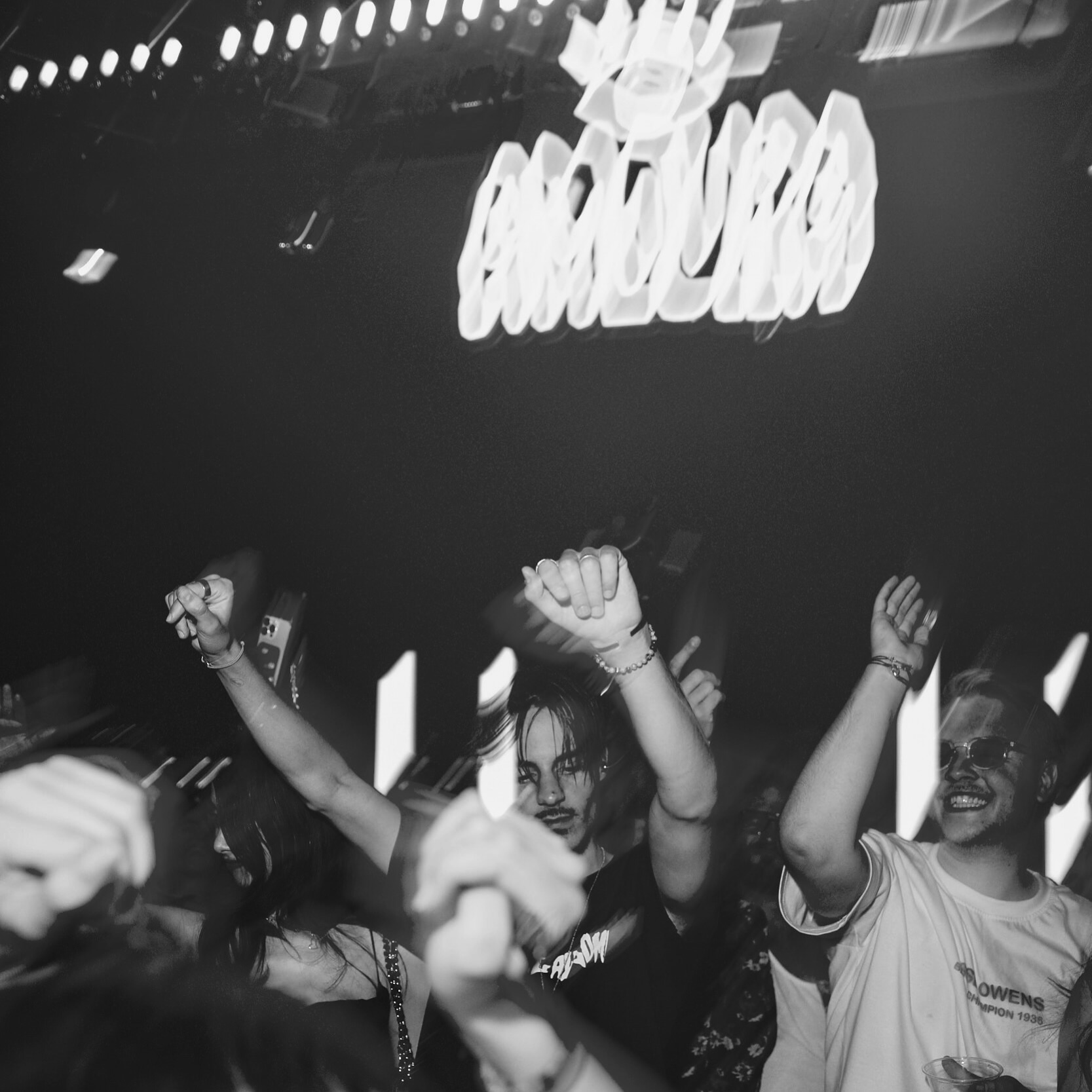 Black and white photo of people dancing energetically in a club, with a blurred sign above reading "Amura.