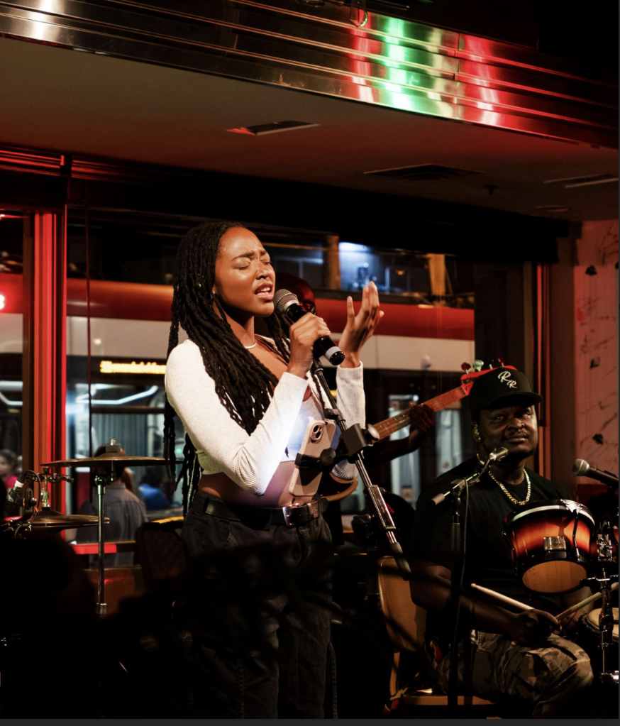 A woman sings passionately into a microphone on stage, while a man next to her plays a drum. The background features red lighting and reflections.