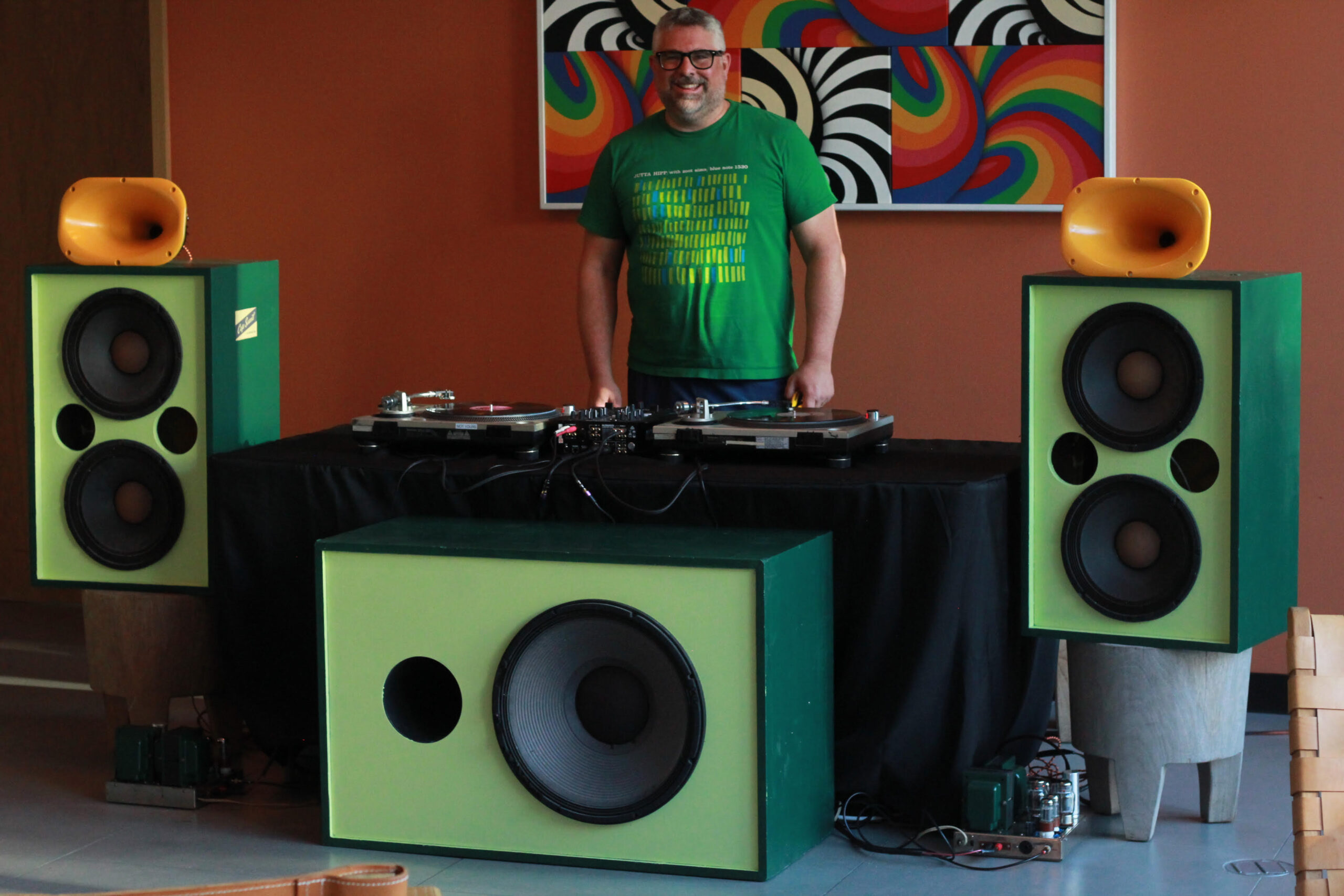 A person stands behind a DJ setup with large green speakers, a turntable, and colorful artwork on the wall.