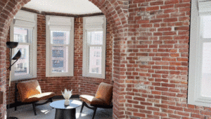 Cozy room with brick walls, featuring two armchairs, a round table with a vase, and a tall lamp. Three windows provide natural light.