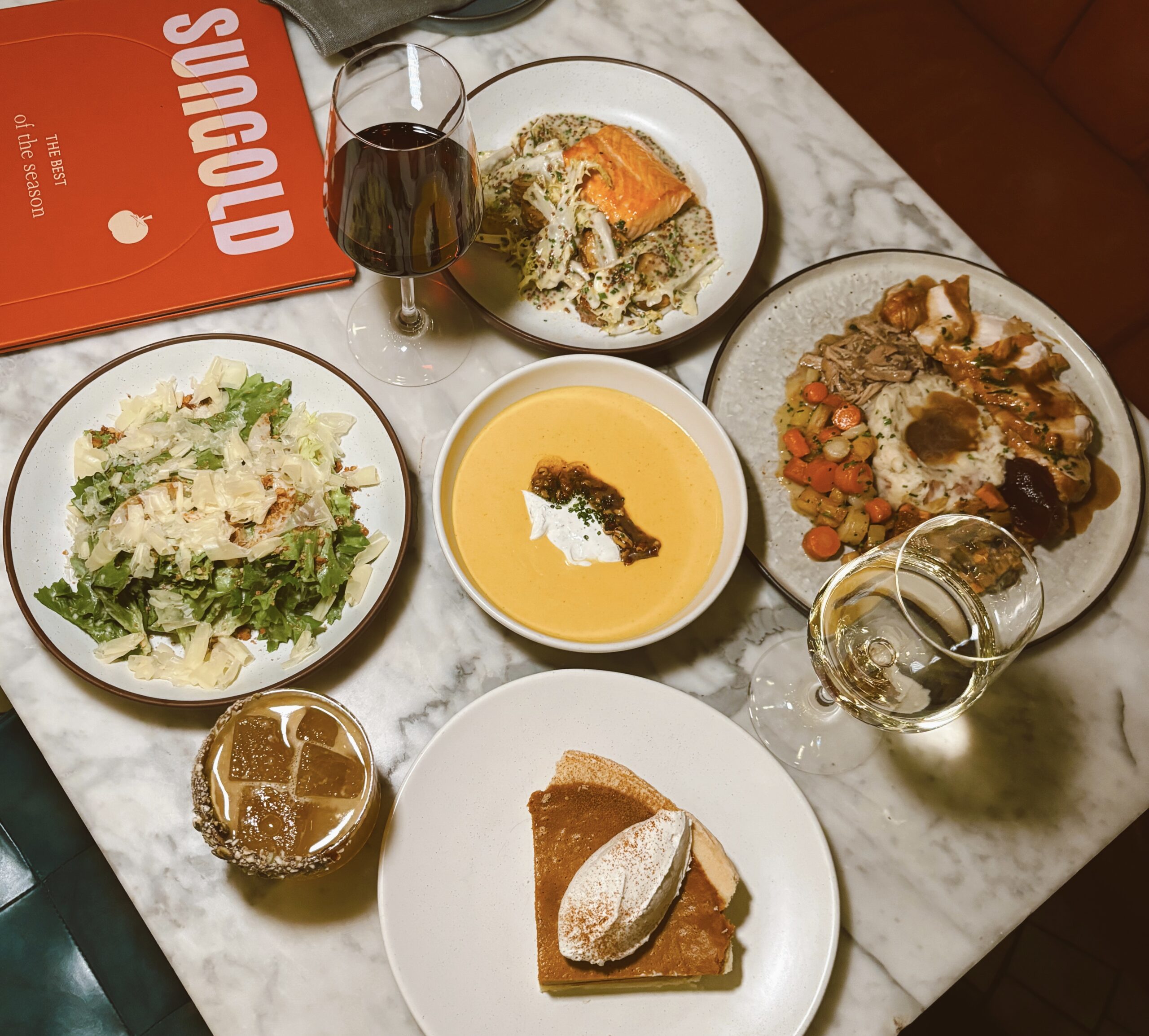 A table with assorted dishes: salad, creamy soup, salmon, meat with vegetables, and a slice of cake. Accompanied by a glass of red and white wine, and a cocktail in a textured glass.