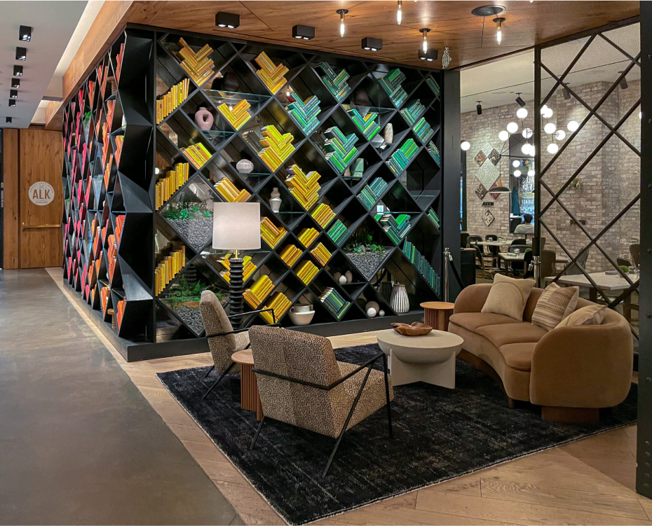 Modern lounge with a colorful diagonal bookshelf, armchairs around a round table, and a sofa on a black rug. Hanging lights illuminate the space, with wooden and glass elements throughout.