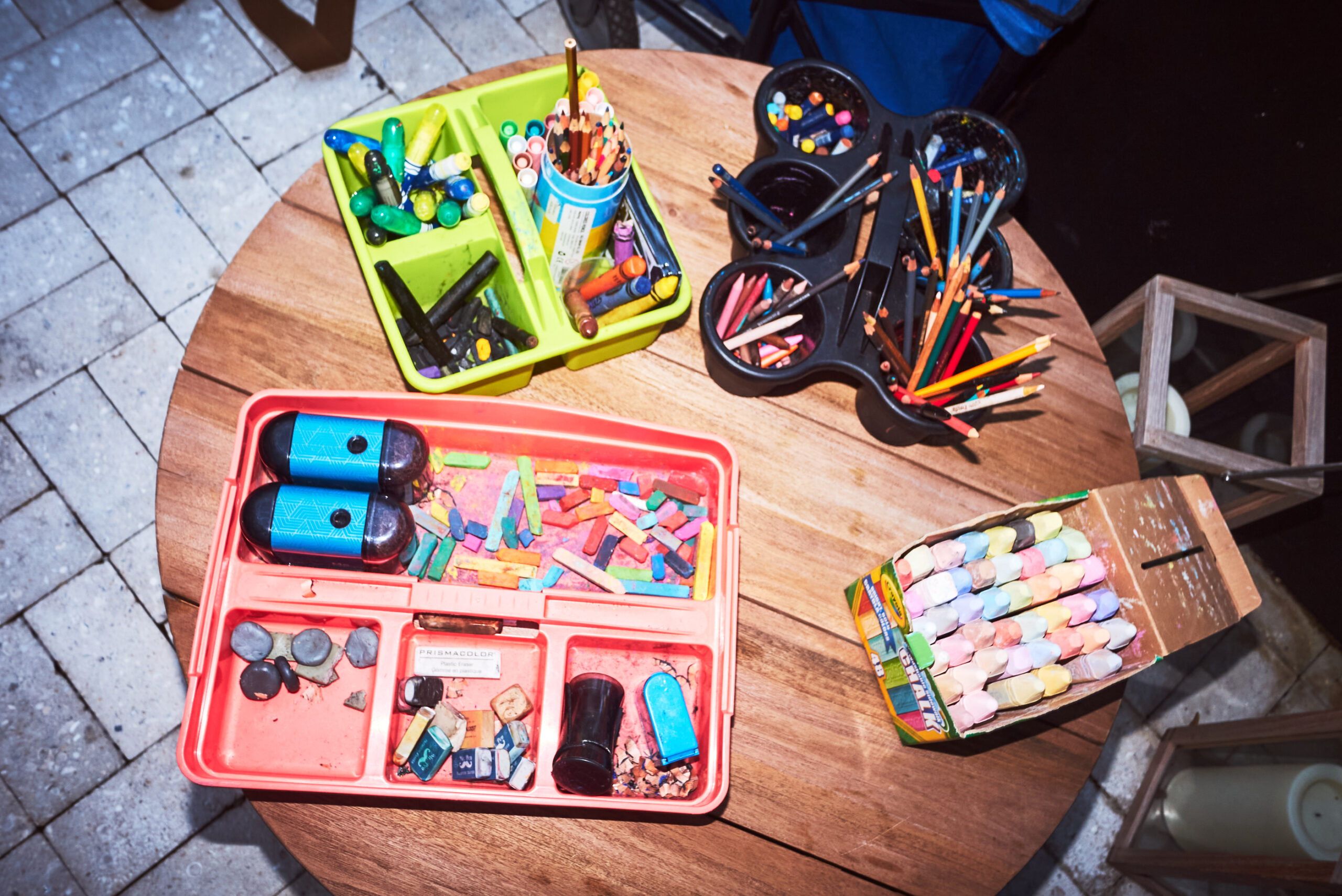 Art supplies including crayons, pastels, pencils, and markers are organized in trays and containers on a round wooden table.