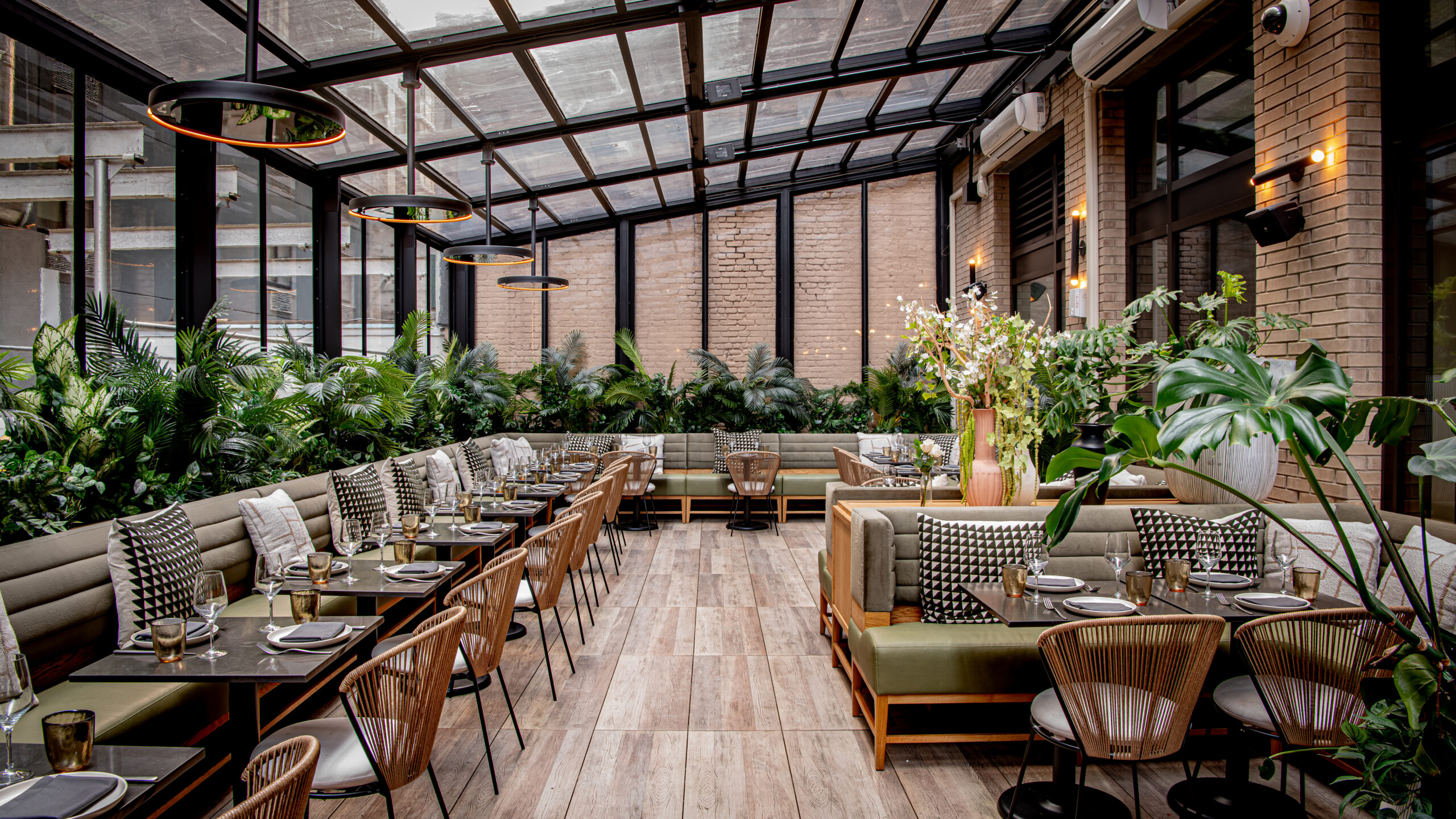 A modern restaurant interior with a glass ceiling, featuring wooden flooring, green plants, and neatly arranged tables and chairs.