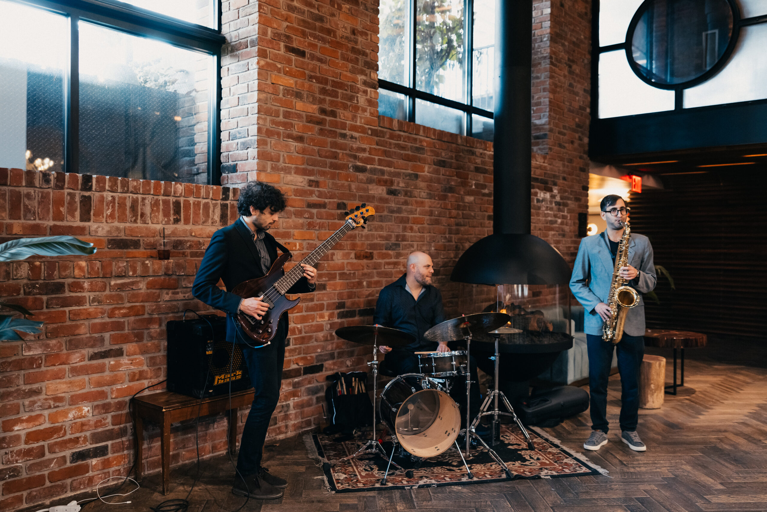 Three musicians perform in a brick-walled venue. A bassist stands on the left, a drummer is seated in the center, and a saxophonist stands on the right.