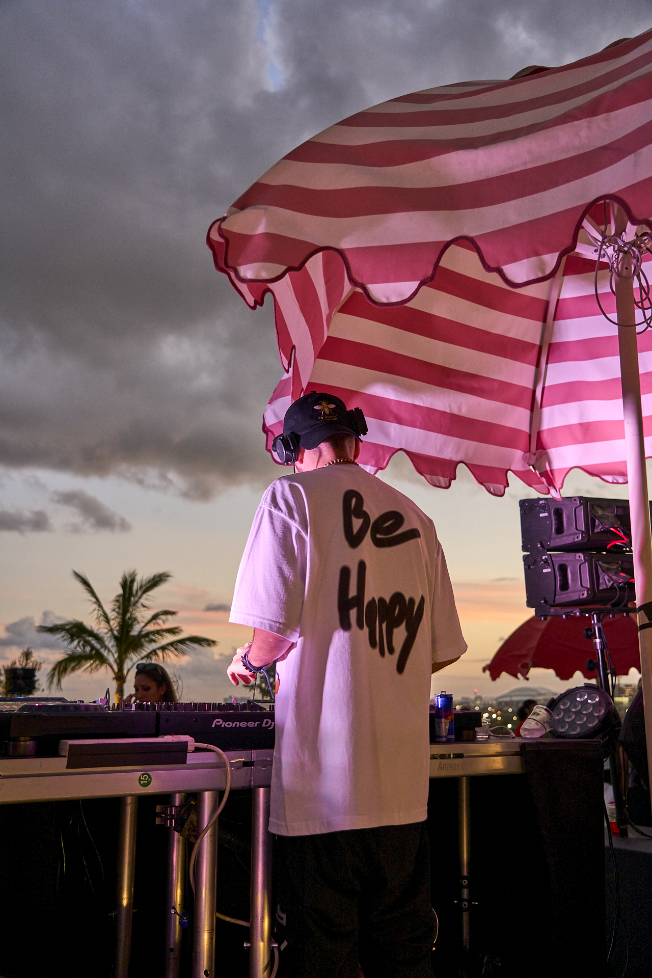 DJ wearing headphones and a "Be Happy" shirt is performing at an outdoor event under a striped umbrella, with a palm tree and cloudy sky in the background.