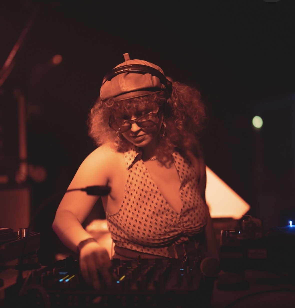 A DJ with curly hair and sunglasses adjusts the settings on a mixer in a dimly lit room.