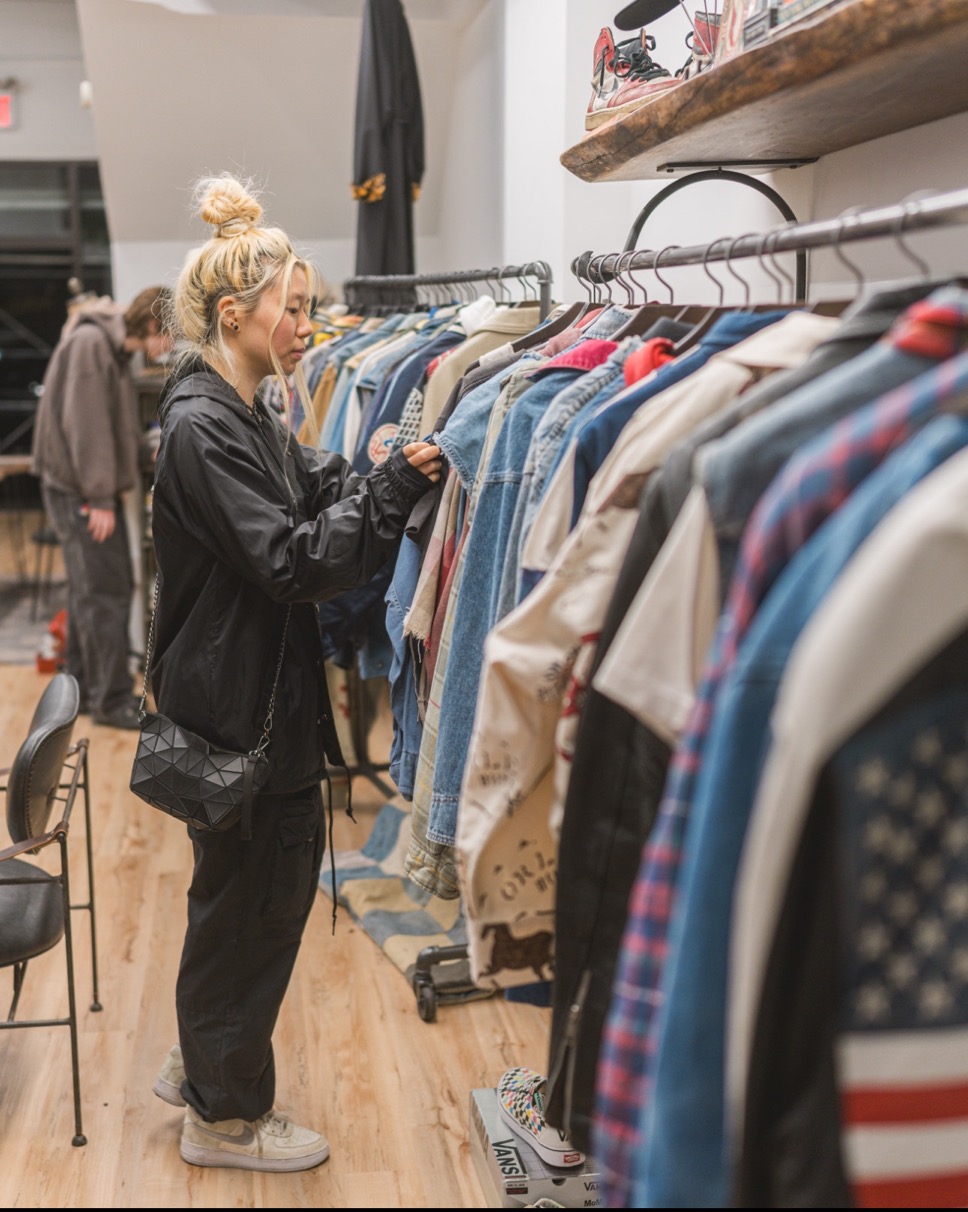 A person with blonde hair browses through a rack of clothes in a store, wearing a black jacket and pants, and carrying a black bag.