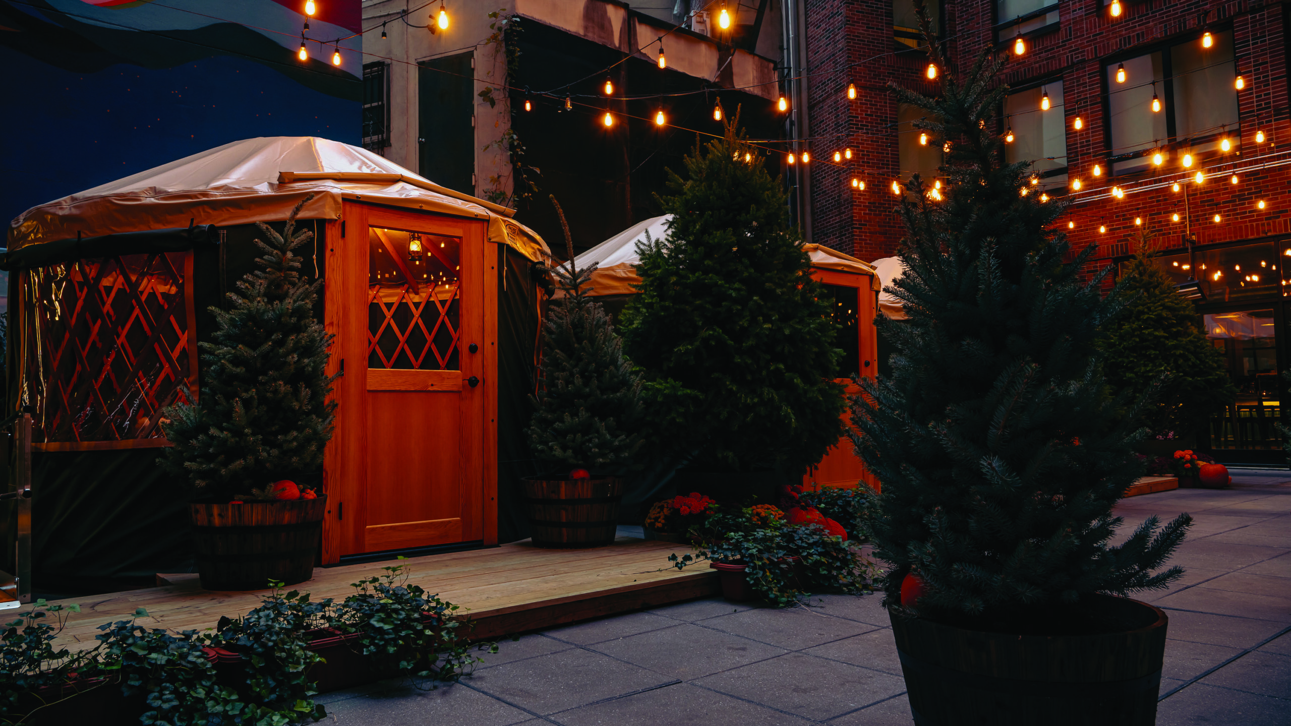 Outdoor seating area with wooden huts, string lights, and potted evergreen trees on a patio.