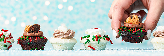 A hand picking up a chocolate cupcake topped with sprinkles and a cookie. Other decorated cupcakes are lined up on a light blue surface with a bokeh background.