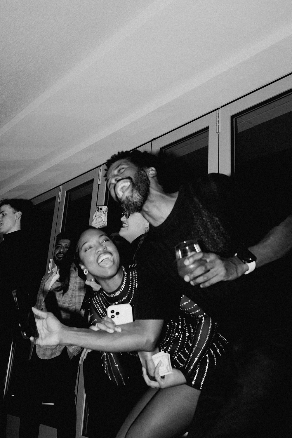 Black and white photo of people enjoying a party. Two people in the foreground are dancing and smiling, one holding a phone, with others talking in the background.