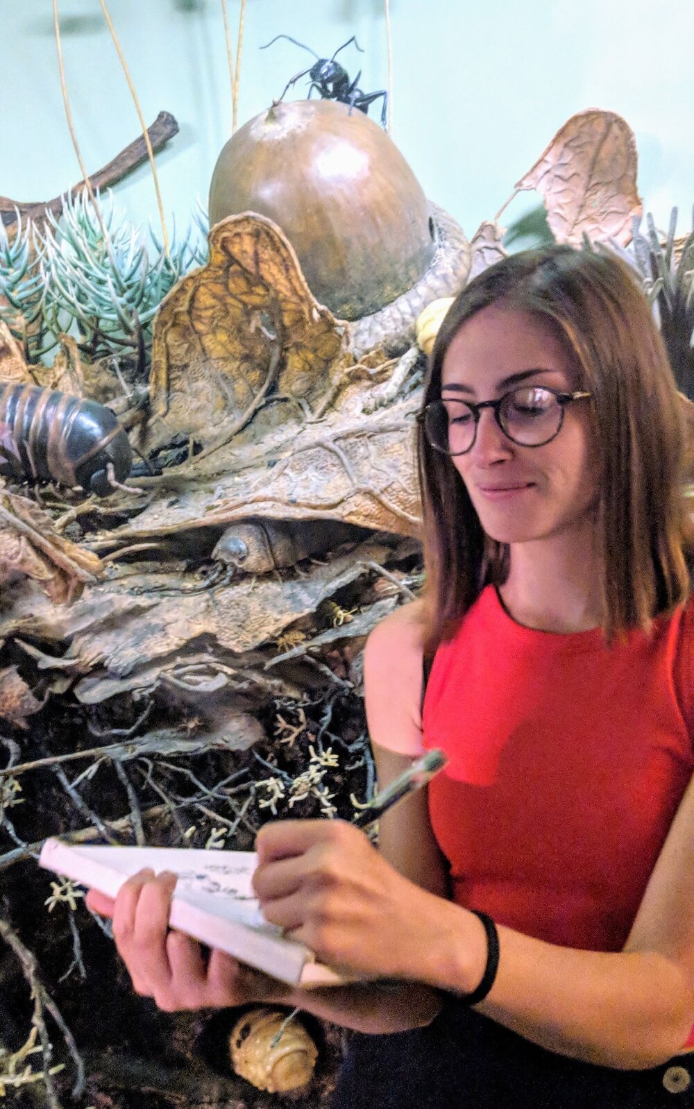 A woman in a red shirt takes notes in front of a display featuring large insects and plants.