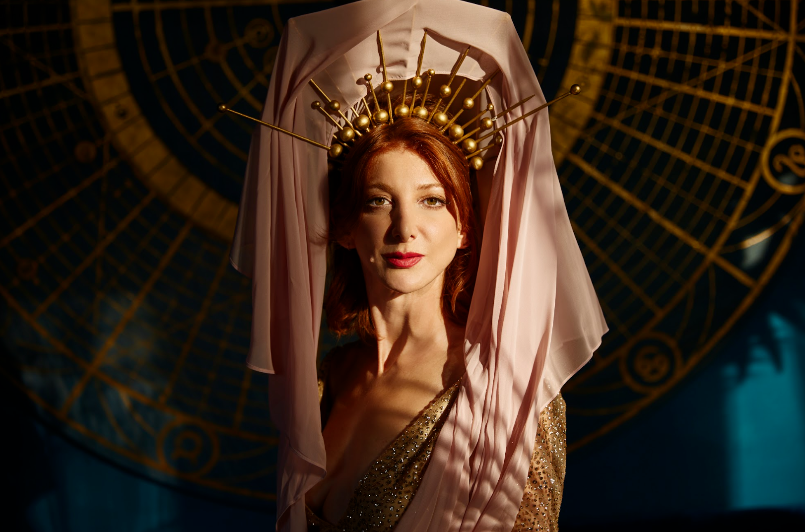 A woman with red hair wears a pink veil and a gold crown made of rods. She stands in front of a dark, geometric circular design.