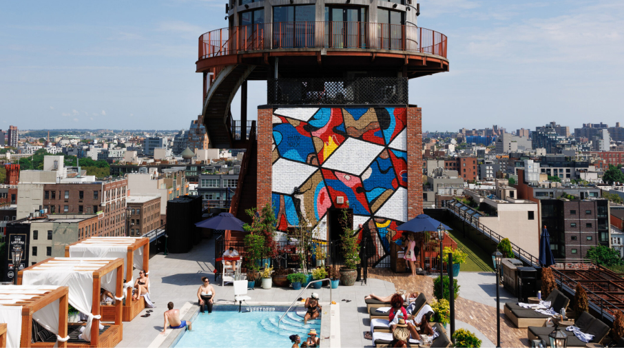 Rooftop pool area with people lounging, a colorful mural on a brick wall, and an old water tower in the background. Urban cityscape visible under a partly cloudy sky.