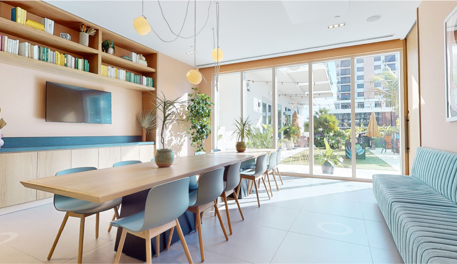 Modern dining area with a long wooden table, multiple chairs, a built-in shelf with books and plants, and large glass doors opening to a sunlit patio with more plants.