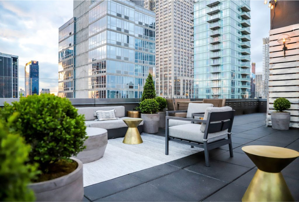 Modern rooftop terrace with seating, potted plants, and gold accent tables, surrounded by city skyscrapers under a partly cloudy sky.