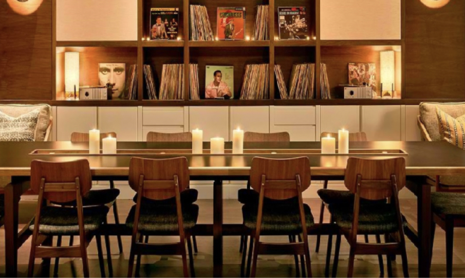 A cozy room with a long dining table, wooden chairs, and lit candles. A bookshelf holds vinyl records and decor items in the background.