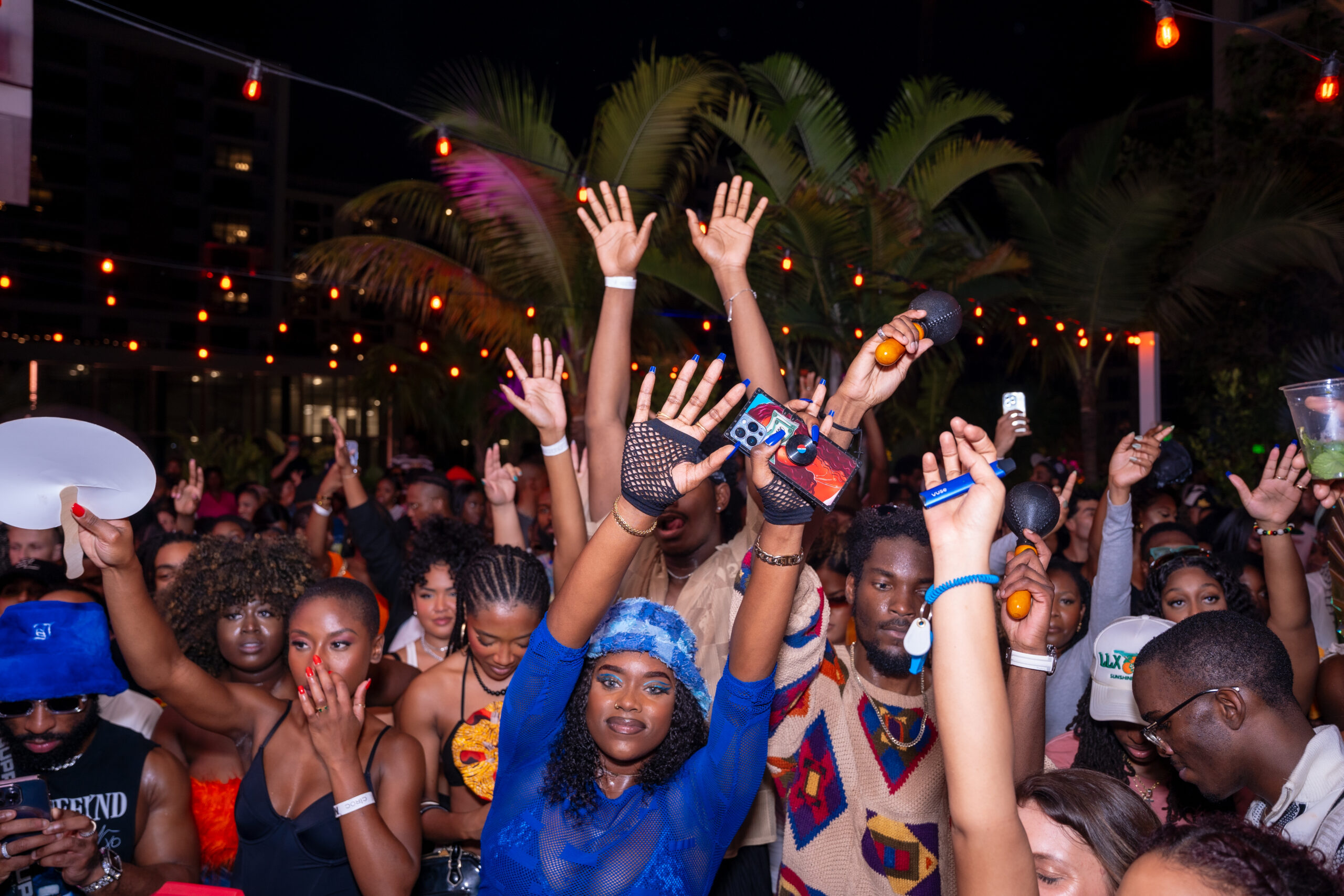 A crowd of people with hands raised, enjoying an outdoor nighttime event. Palm trees and string lights are visible in the background.