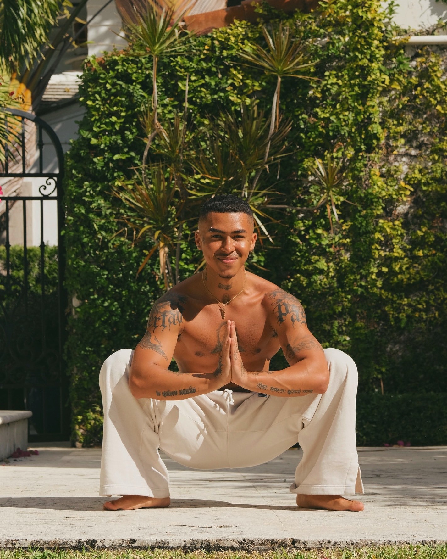 Person with tattoos squatting outdoors, hands in prayer position, wearing white pants. Background features lush greenery and a decorative gate.