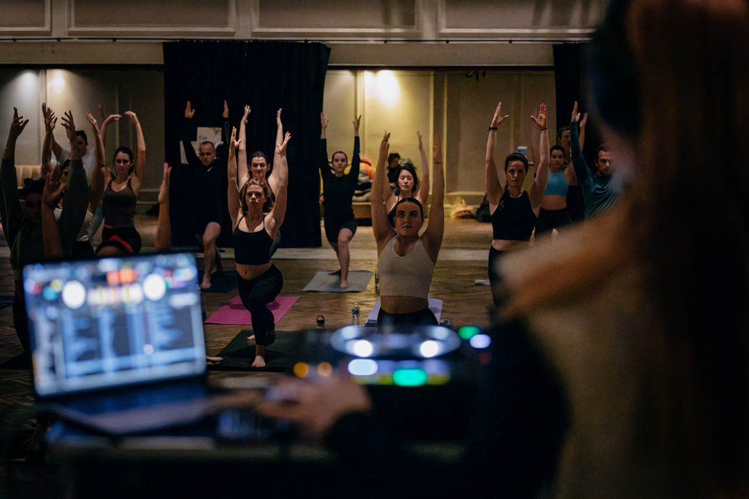 A group of people performing yoga poses in a dimly lit room, with a DJ booth and equipment visible in the foreground.
