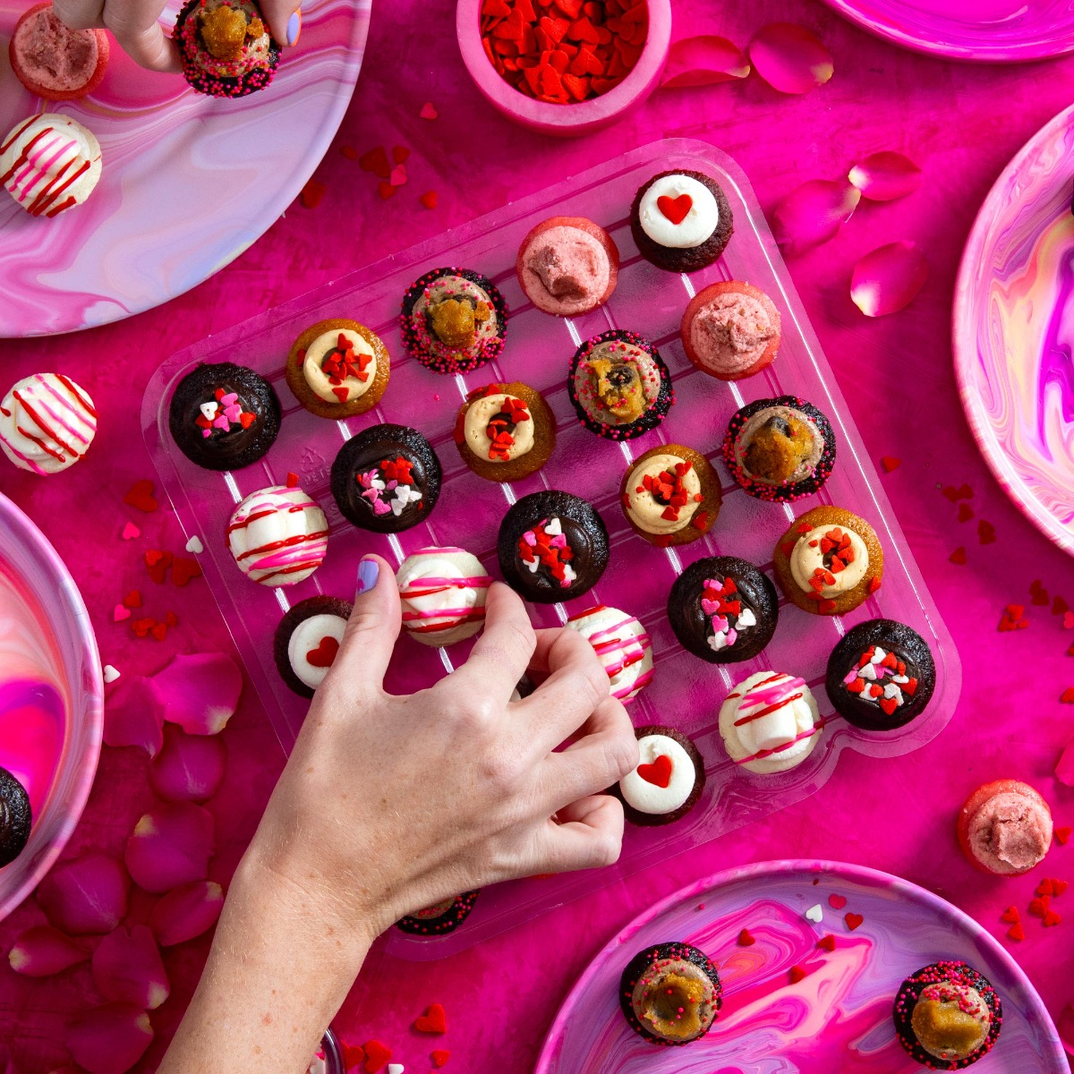 A hand reaches for a mini cupcake from a tray on a pink marbled table. The cupcakes are decorated with various toppings like hearts and sprinkles.