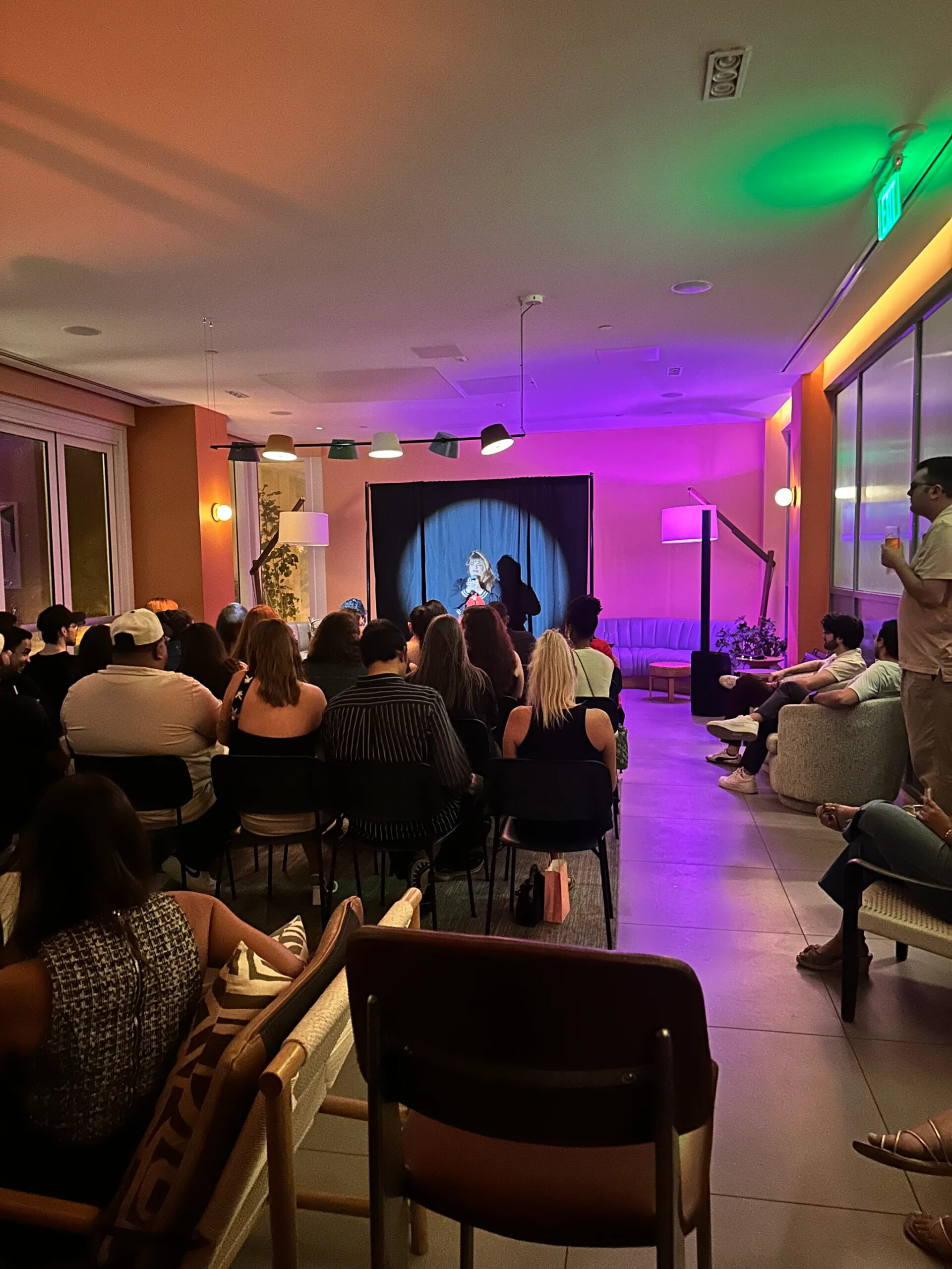 A comedian performs on a small stage in front of an audience seated in a dimly lit room.