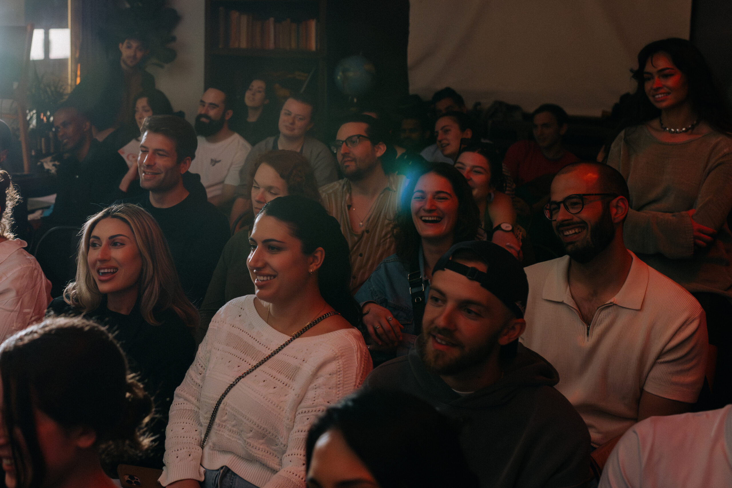 A group of people seated closely together indoors, attentively watching something, with several individuals smiling and laughing.