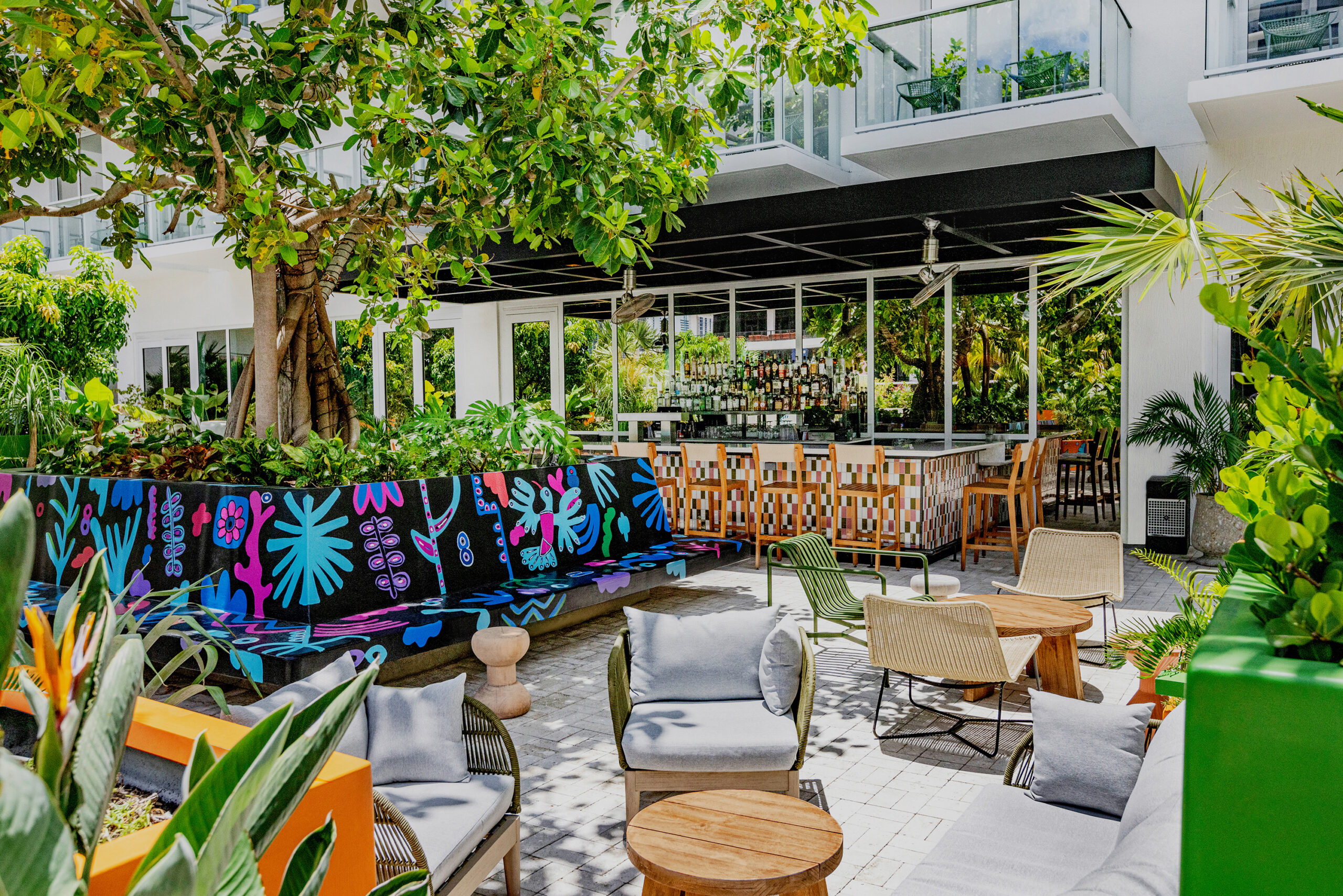 Outdoor seating area with modern furniture, a colorful mural, and lush greenery. A bar with stools is in the background under a canopy.